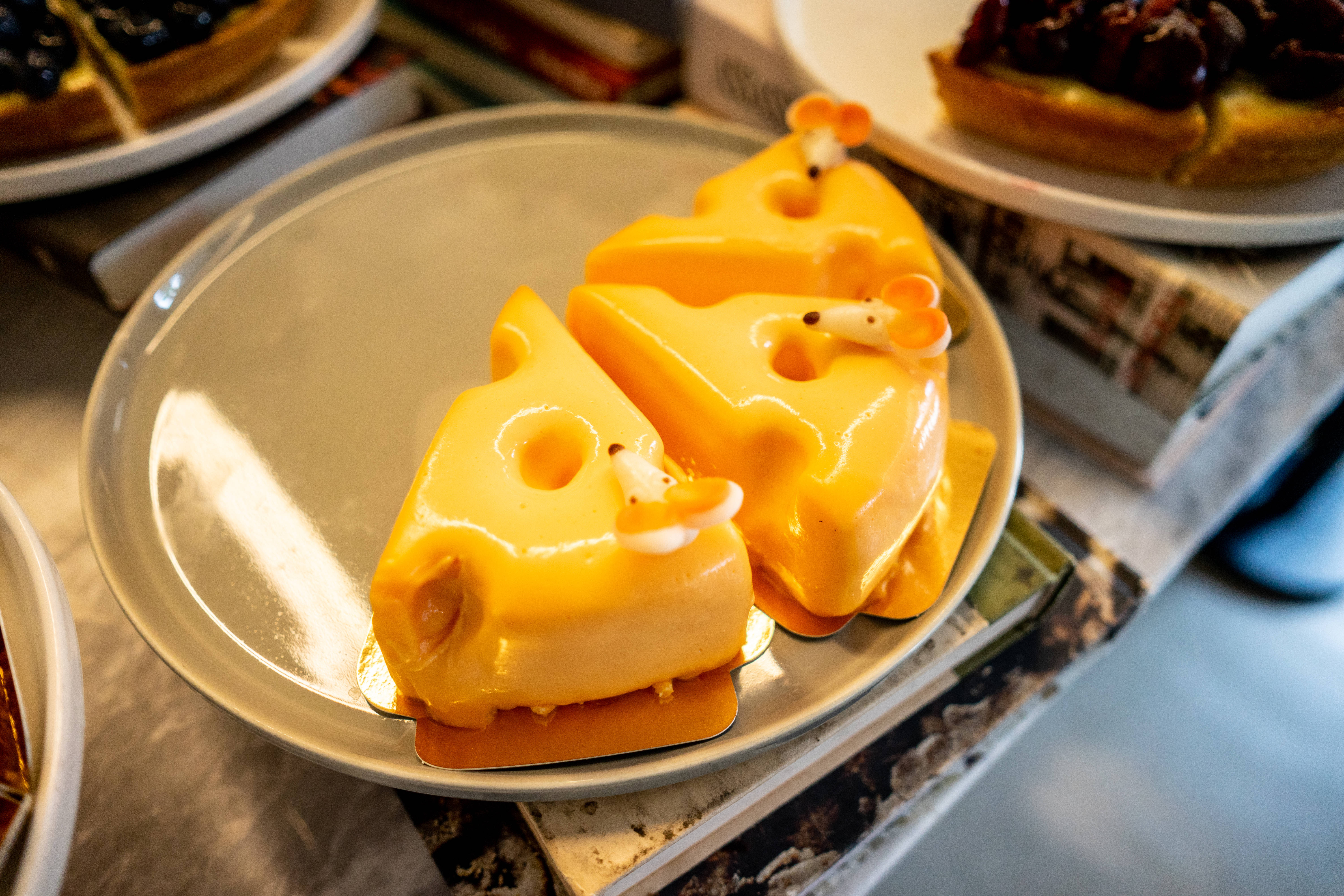 Cheese 桌布 壁紙 — 兩個光澤黃色起司形狀的糕點，帶有細小的老鼠裝飾，放在銀色盤子上，置於疊起的雜誌上。