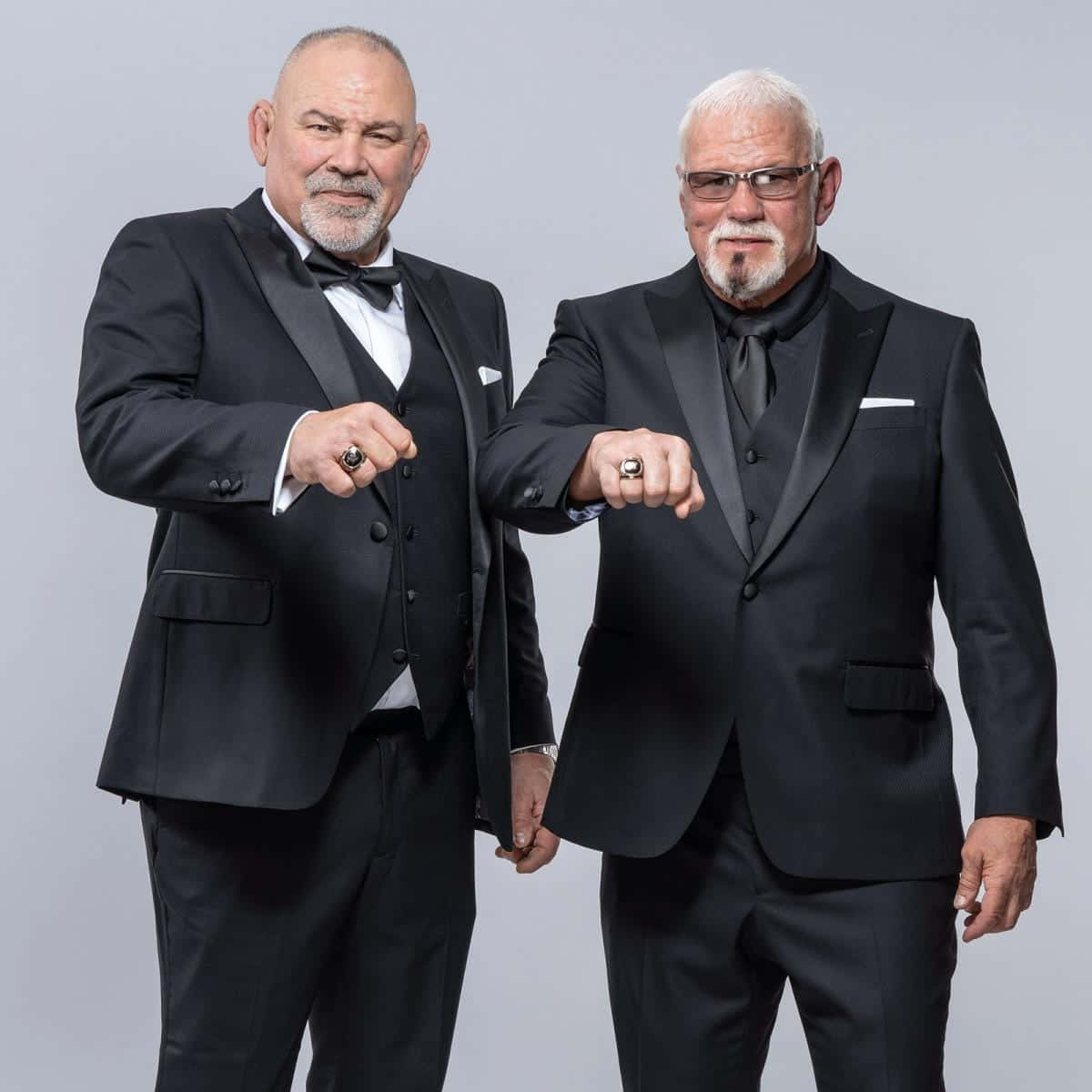 Rick Steiner 桌布 壁紙 — 兩個光頭男人穿著黑色燕尾服，在純灰色背景前自信地舉起拳頭擺姿勢。