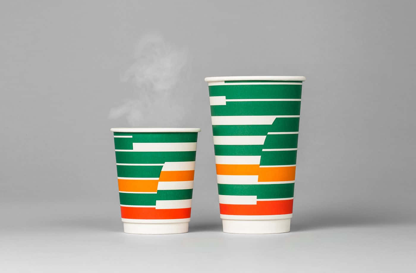 7-ELEVEN 桌布 壁紙 — 兩個冒著蒸氣的7-Eleven紙杯，帶有綠色、橙色和白色條紋，並排放在中性灰色表面上。