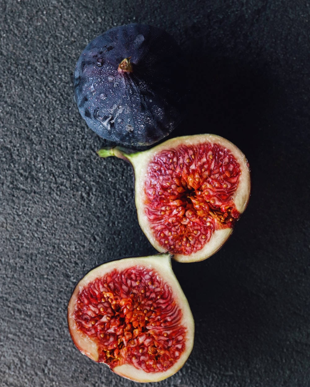 Fruit 桌布 壁紙 — 兩個切半的成熟無花果，帶有鮮紅色的果肉，和一個完整的深色無花果，放在有紋理的黑色表面上。