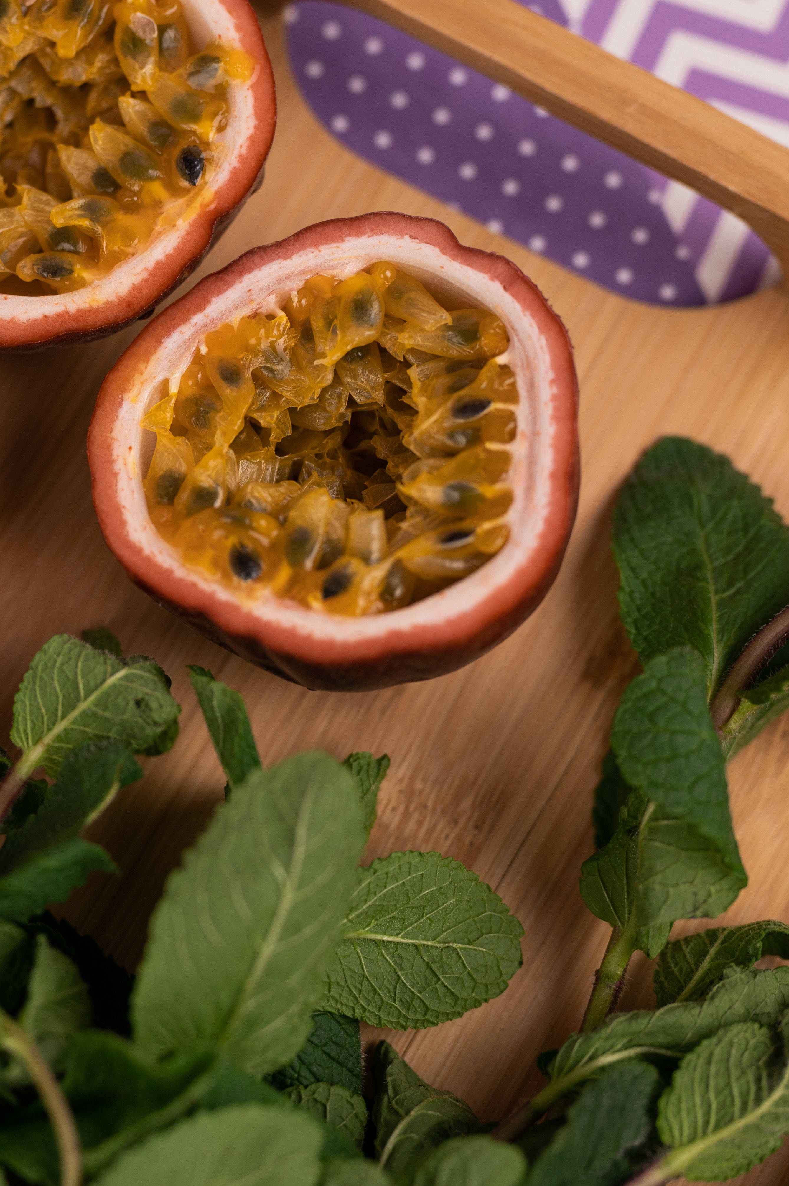 Fruit 桌布 壁紙 — 兩個切半的百香果，帶有多汁的黃色果肉和黑色種子，放在木板上，旁邊是新鮮薄荷葉和紫色圖案布。