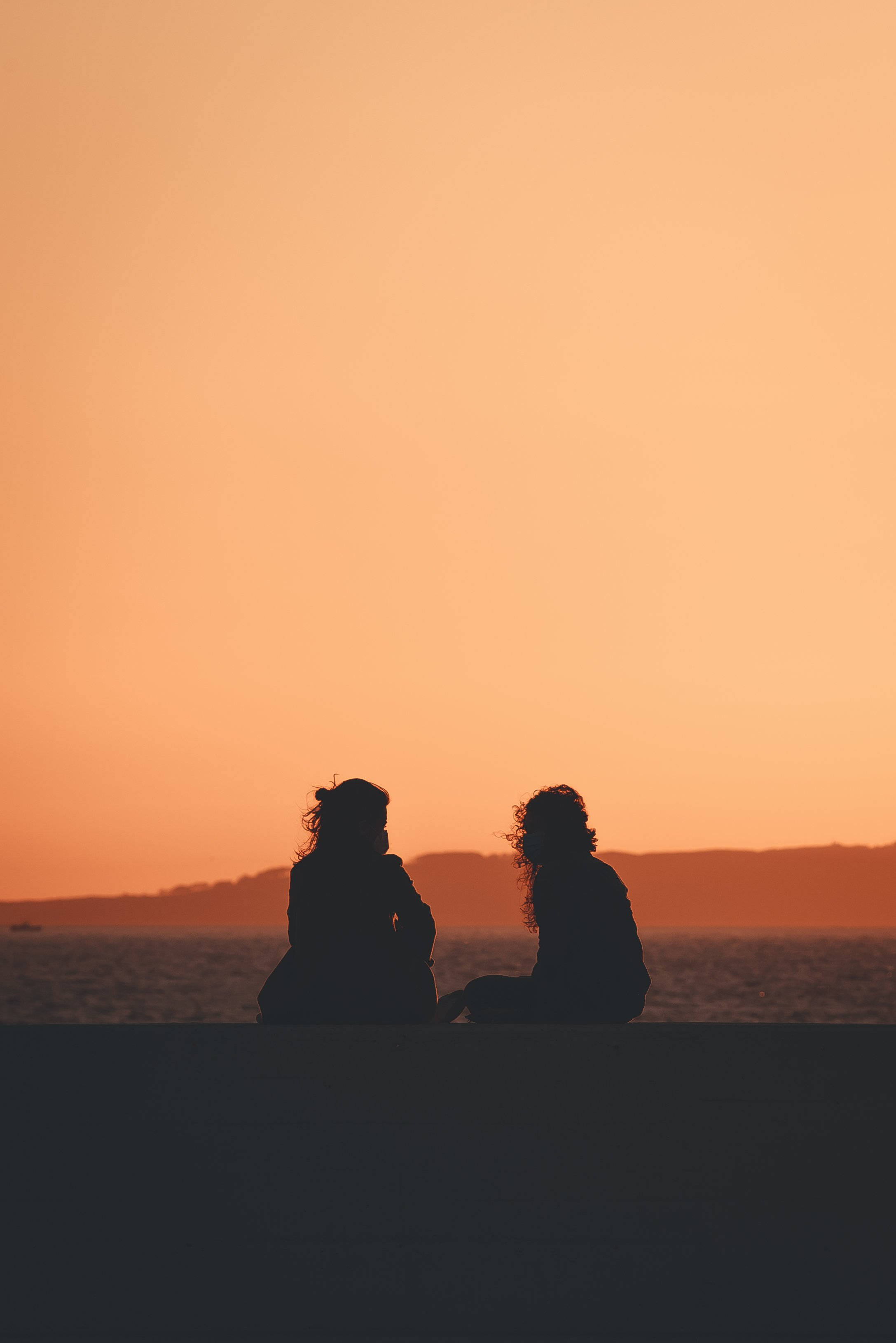 Silhouette 桌布 壁紙 — 兩個剪影人物並排坐在碼頭上，夕陽以溫暖的橙色調在水面上落下。