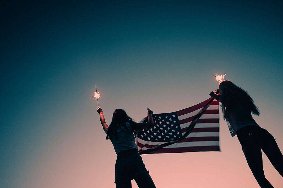 Freedom 桌布 壁紙 — 兩個剪影人物在帶有藍色和粉紅色漸變的暮色天空中手持美國國旗和煙火棒。