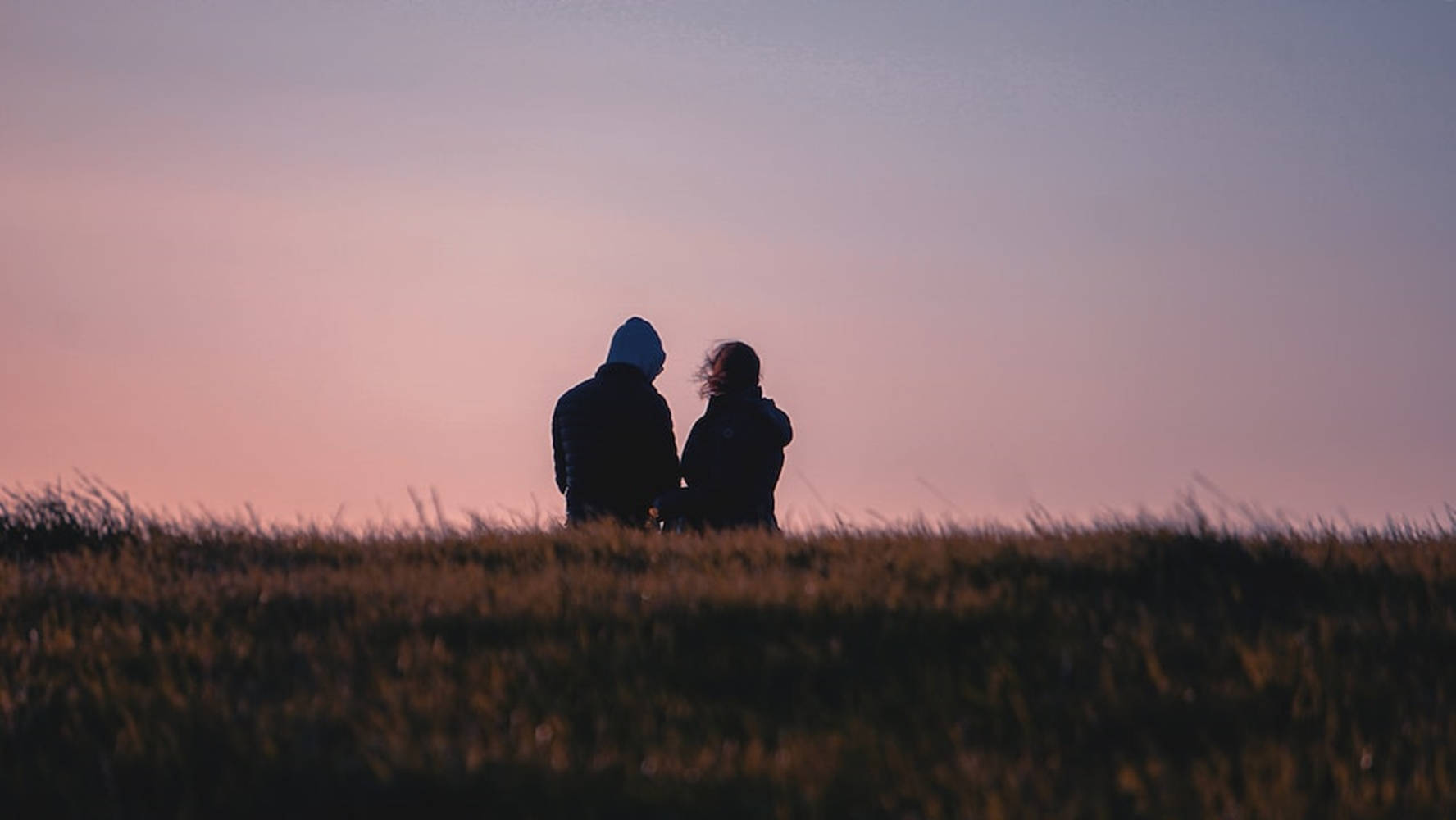 couple love 桌布 壁紙 — 兩個剪影人物在日落時分緊密地站在長滿草的山丘上，沐浴在柔和的粉紅色和紫色暮光中。