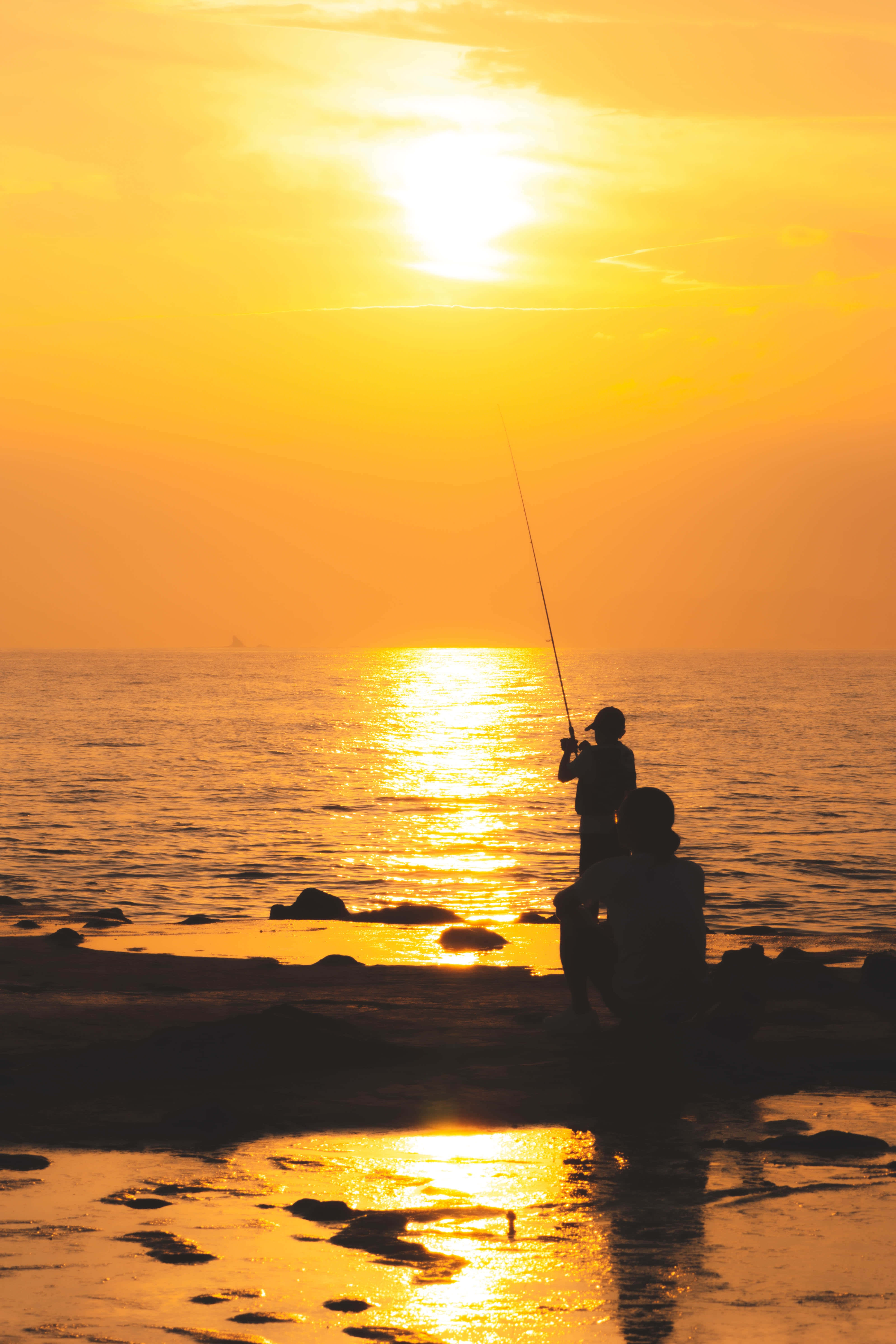 Fishing Phone 桌布 壁紙 — 兩個剪影人物在日落時分釣魚，金色光線映照在平靜的海水和濕漉漉的岩石上。