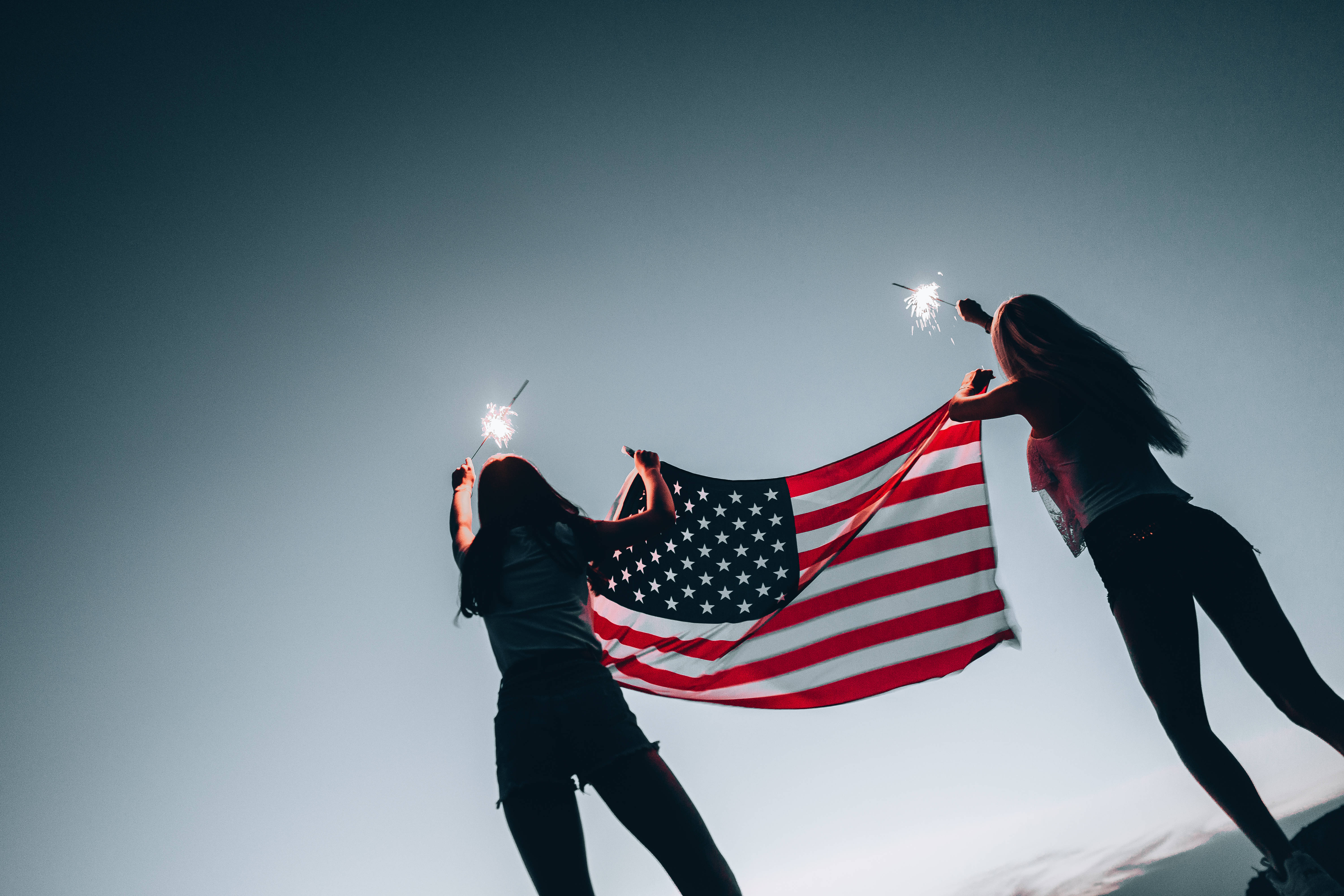 USA flag iPhone 桌布 壁紙 — 兩個剪影人物在暮色天空下手持美國國旗和煙火棒。