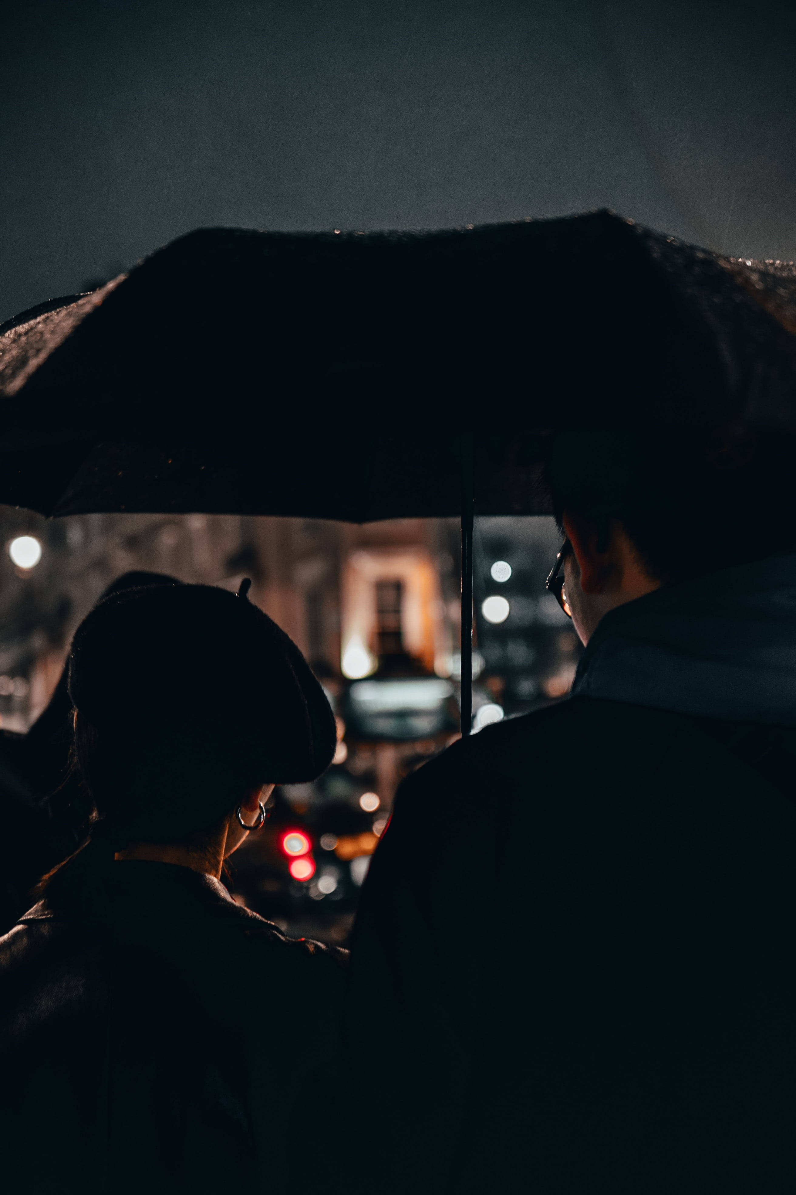情侶 桌布 壁紙 — 兩個剪影人物在雨中共享一把黑色雨傘，背後是夜間模糊的城市燈光。