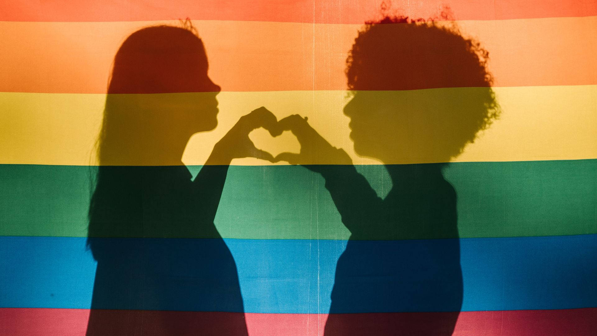Subtle Lgbt 桌布 壁紙 — 兩個剪影人物在鮮豔的彩虹旗背景下用手比出心形。
