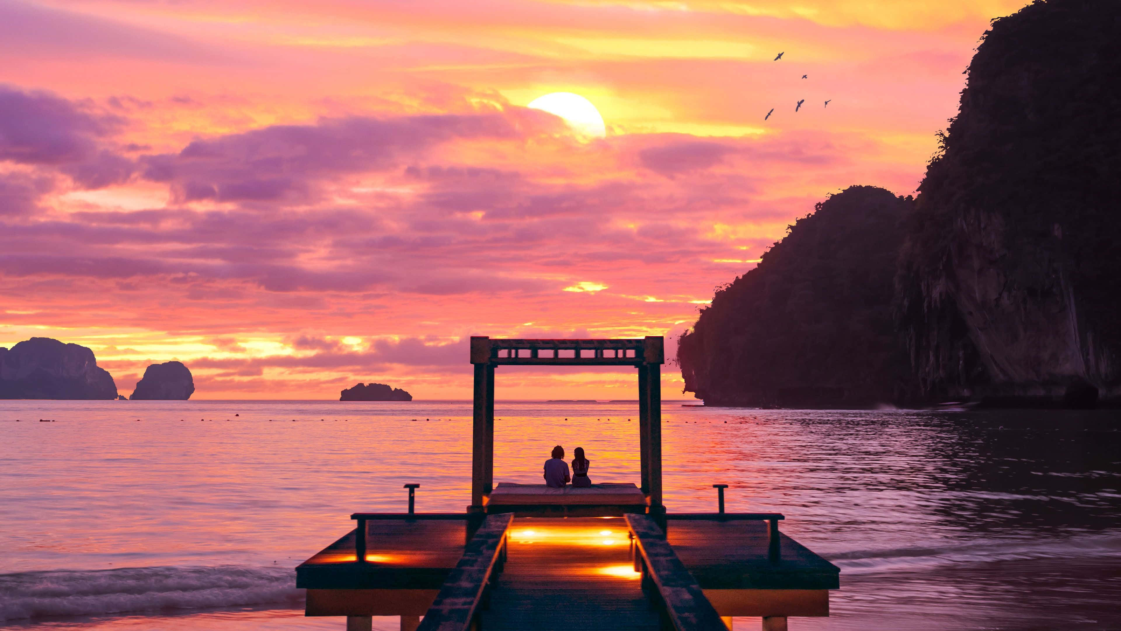 Sweet Love 桌布 壁紙 — 兩個剪影人物坐在日落時分發光的木碼頭上，以平靜的海洋和岩石懸崖上充滿活力的粉紅色和橙色雲彩為框架。