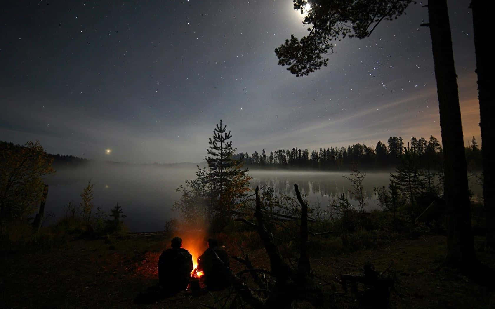 Campfire 桌布 壁紙 — 兩個剪影人物坐在發光的營火旁，上方是繁星點點的夜空，一彎新月懸在霧氣瀰漫的湖面上。
