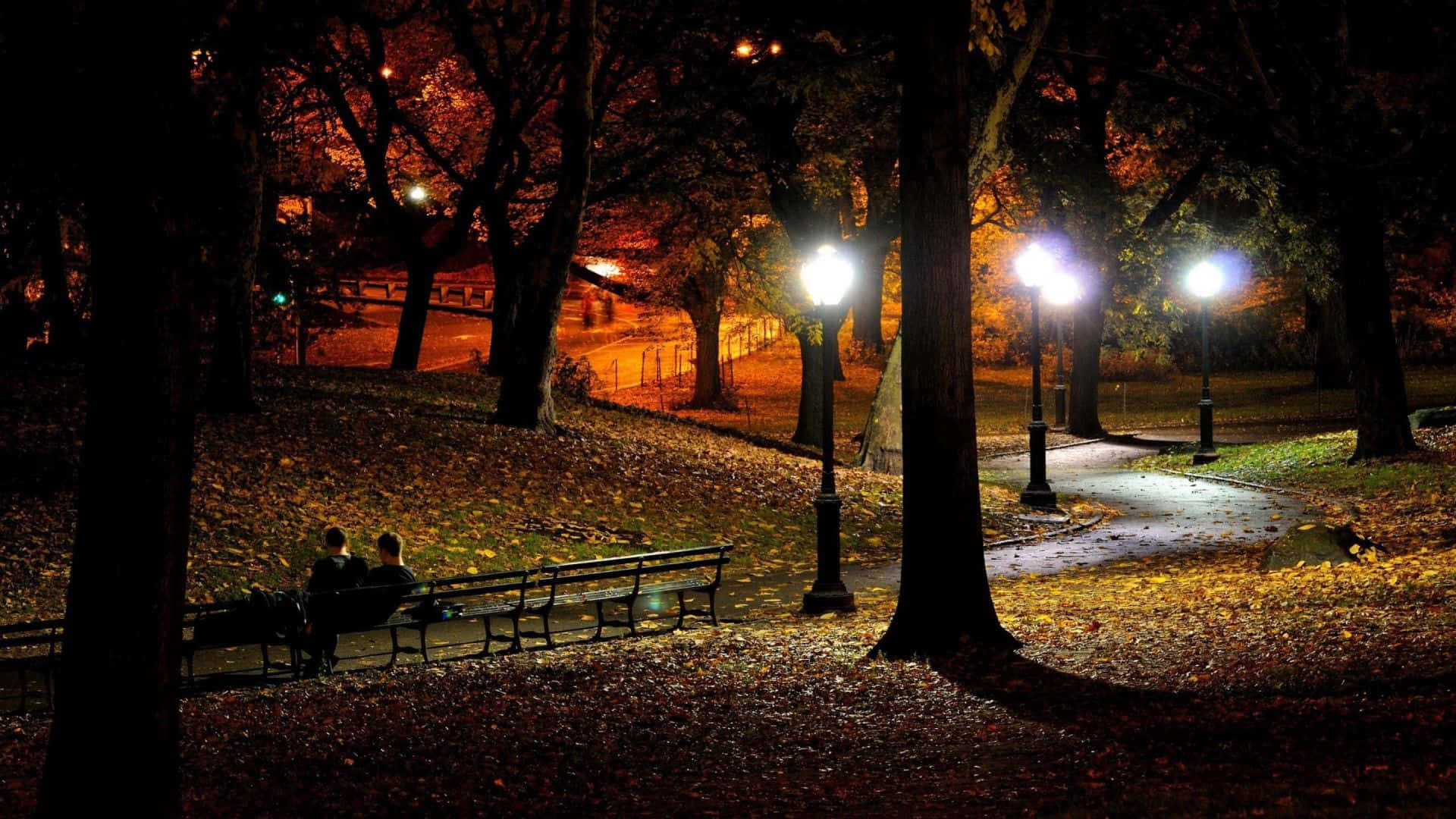 Autumn Evening 桌布 壁紙 — 兩個剪影人物坐在長椅上，在發光的街燈下，周圍是寧靜公園夜間鋪滿的落葉地毯。