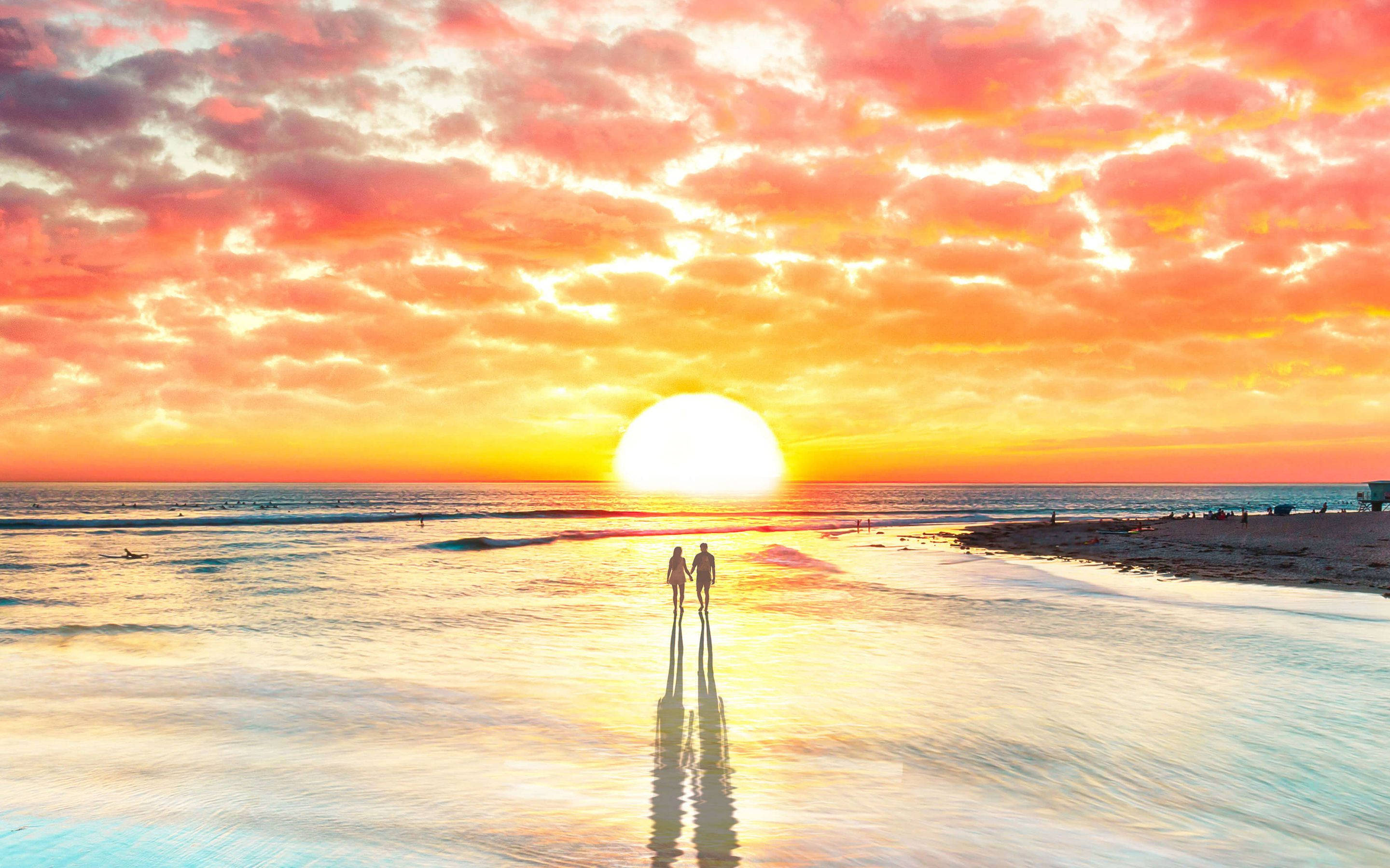 情侶 桌布 壁紙 — 兩個剪影人物手牽手沿著濕漉漉的海灘漫步，夕陽在火紅的橙色和粉紅色天空中落下。
