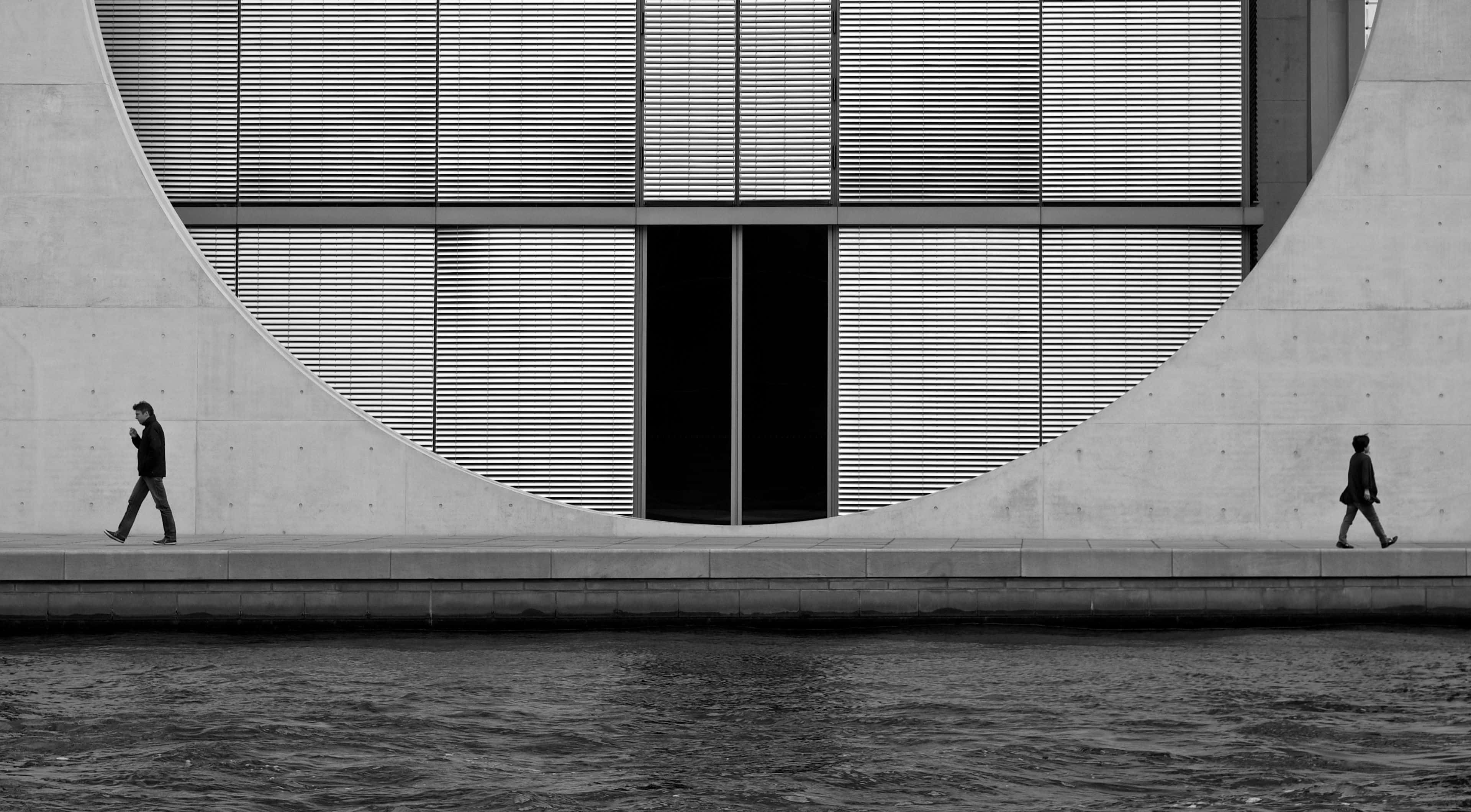 Black And White Architecture 桌布 壁紙 — 兩個剪影人物沿著海濱小徑行走，上方是帶有水平板條的巨大彎曲混凝土拱門。