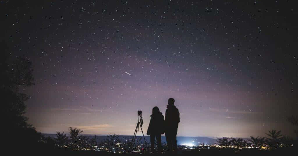 Stars 桌布 壁紙 — 兩個剪影人物站在望遠鏡旁，凝視著繁星點點的夜空，一道微弱的流星劃過頭頂。