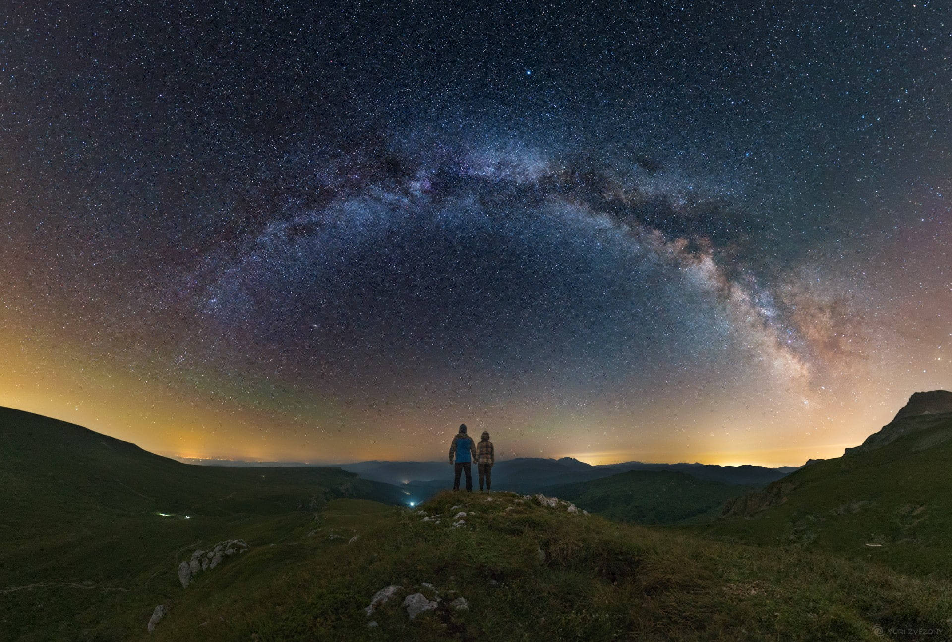 愛心 桌布 壁紙 — 兩個剪影人物站在長滿草的山脊上，仰望橫跨星空、令人嘆為觀止的銀河。