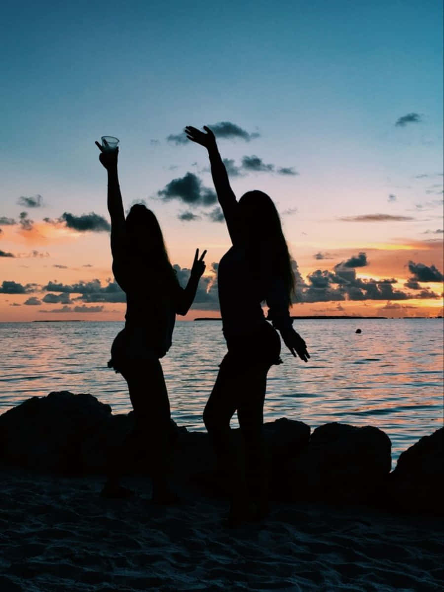 Friends 桌布 壁紙 — 兩個剪影女性在海灘上慶祝日落，舉起手臂和飲料，背景是充滿活力的橙色和藍色天空。