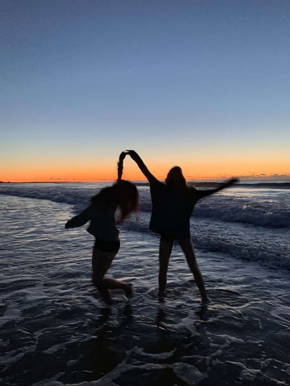 Best Friend Aesthetic 桌布 壁紙 — 兩個剪影朋友在海浪中歡快地跳舞，天空在日落時閃耀著橙色和藍色。