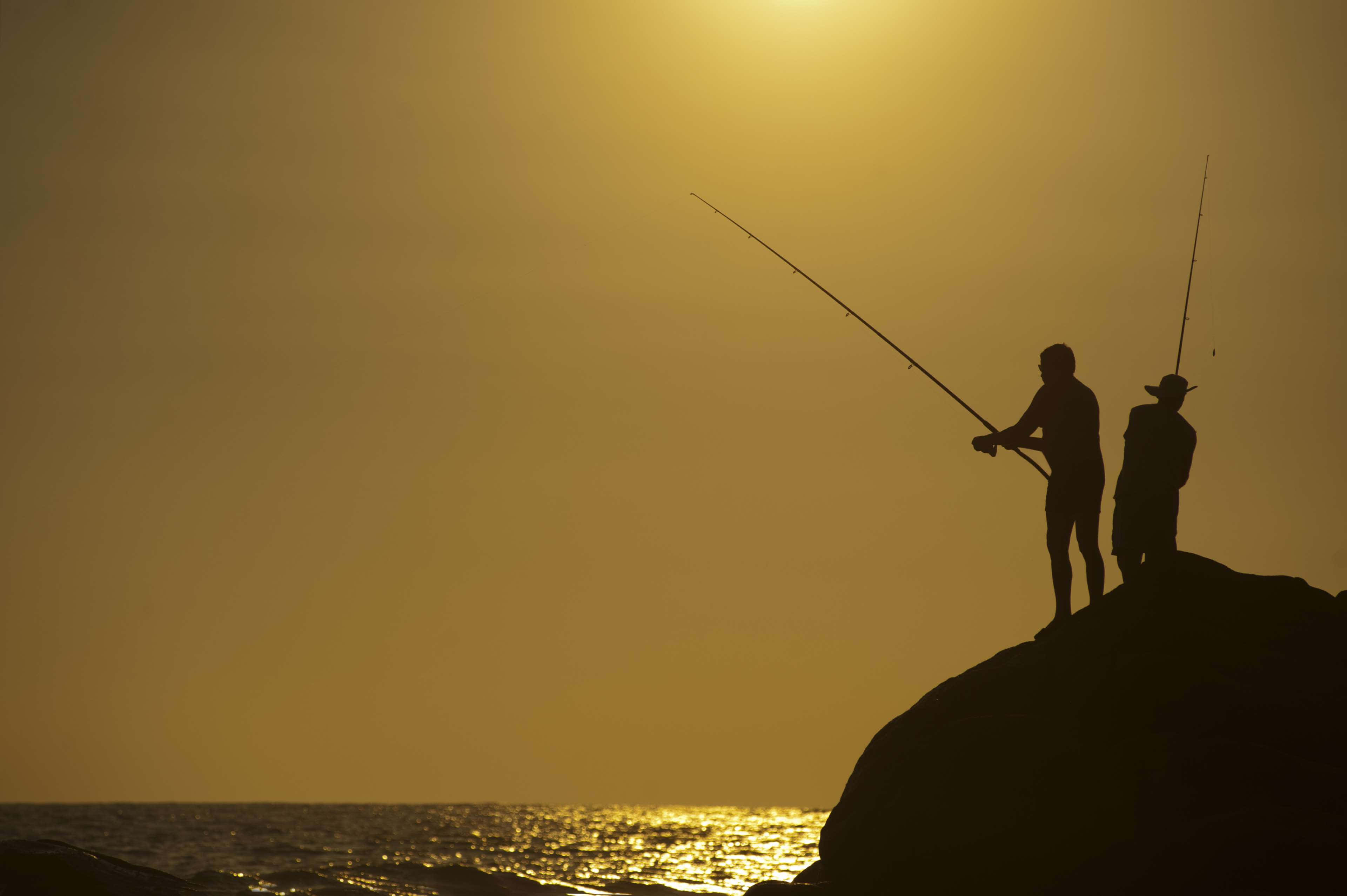 Fishing Rod 桌布 壁紙 — 兩個剪影漁夫站在岩石懸崖上，在金色迷霧中的日落時分將魚線拋入海洋。