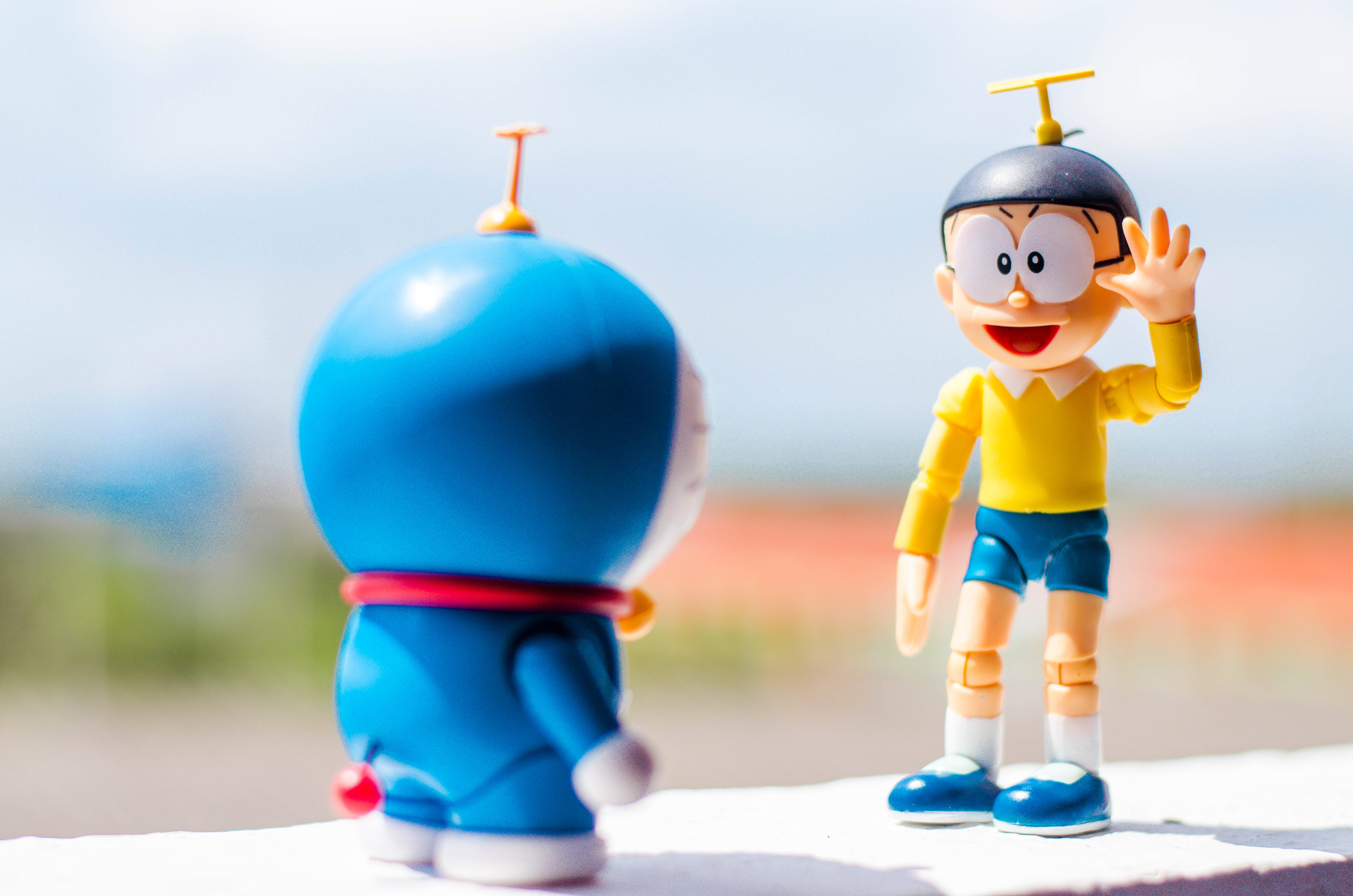 Doraemon 桌布 壁紙 — 兩個動漫人物哆啦A夢和大雄在明亮的日光下，站在一個壁架上，面對面，背景是模糊的戶外景色。