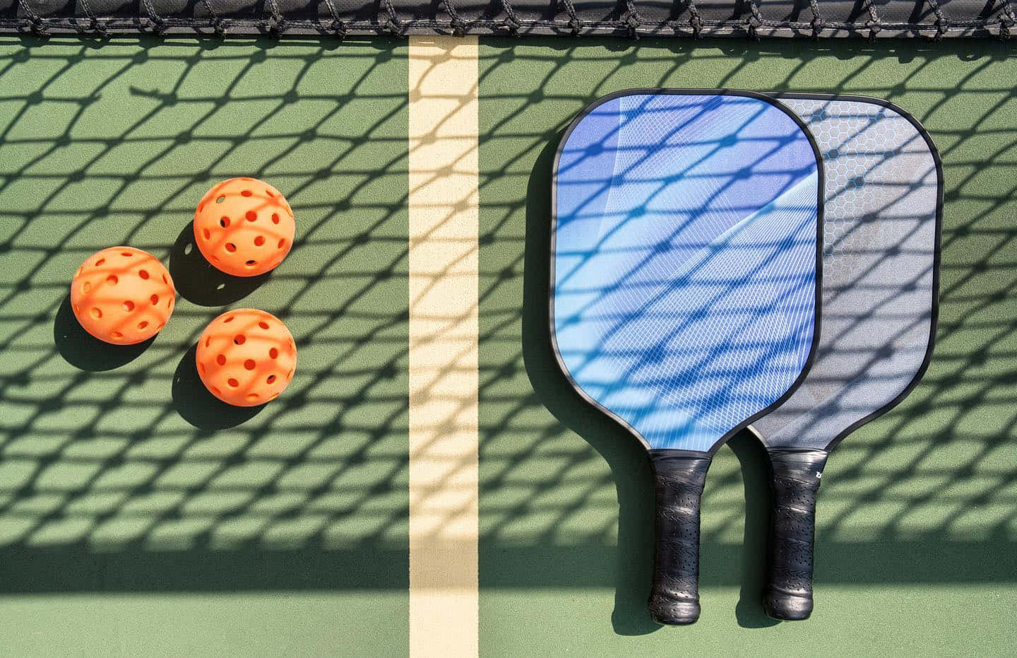 Pickleball 桌布 壁紙 — 兩個匹克球拍和三個橙色球放在綠色球場上，網下投射出清晰的陰影。