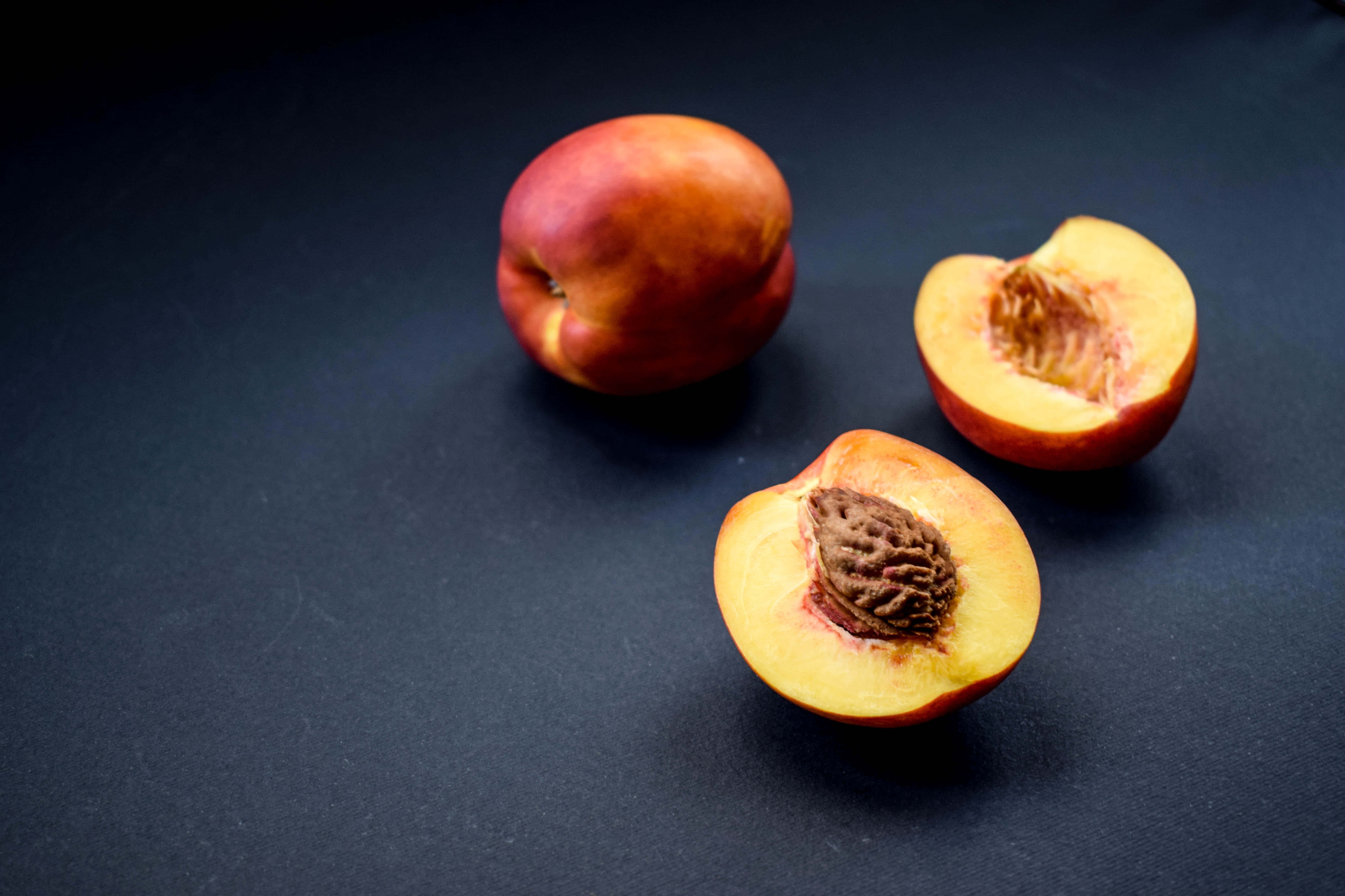 Peaches 桌布 壁紙 — 兩個半邊桃子和一個完整桃子放在深色紋理表面上，展現多汁果肉和絨毛表皮。