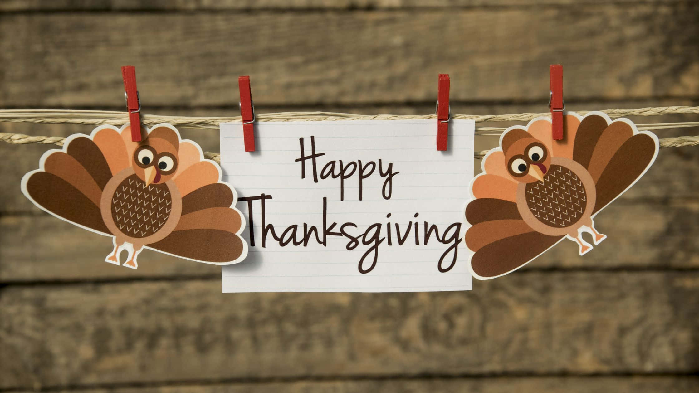 Thanksgiving Day 桌布 壁紙 — 兩個卡通火雞剪紙和一張手寫便條用紅色衣夾掛在繩子上，背景是質樸的木質背景。