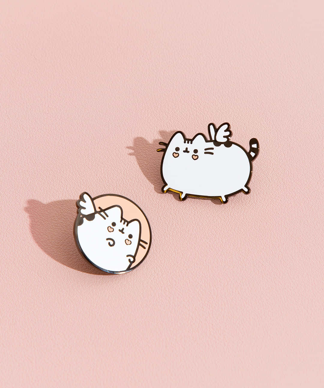 pusheen 動圖 桌布 壁紙 — 兩個可愛的Pusheen琺瑯別針，帶有天使翅膀，放在柔軟的粉紅色紋理表面上，投下微妙的陰影。