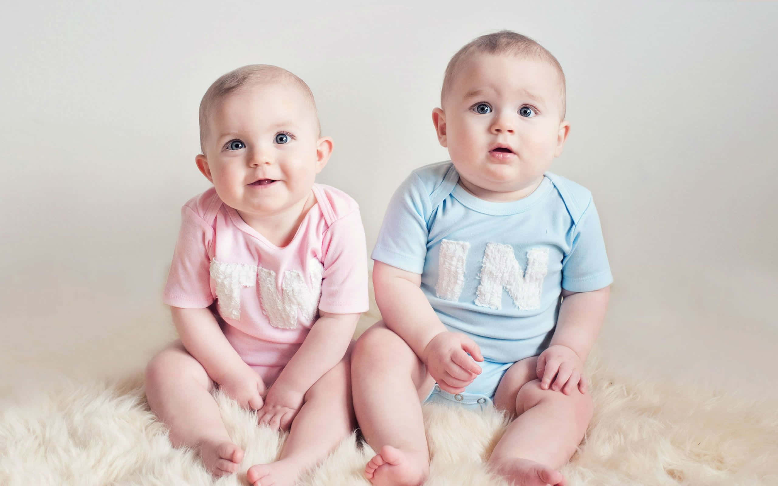 Cute Babies 桌布 壁紙 — 兩個可愛的寶寶並排坐在一張毛茸茸的白色地毯上，一個穿粉紅色，另一個穿藍色，兩人都好奇地看著鏡頭。
