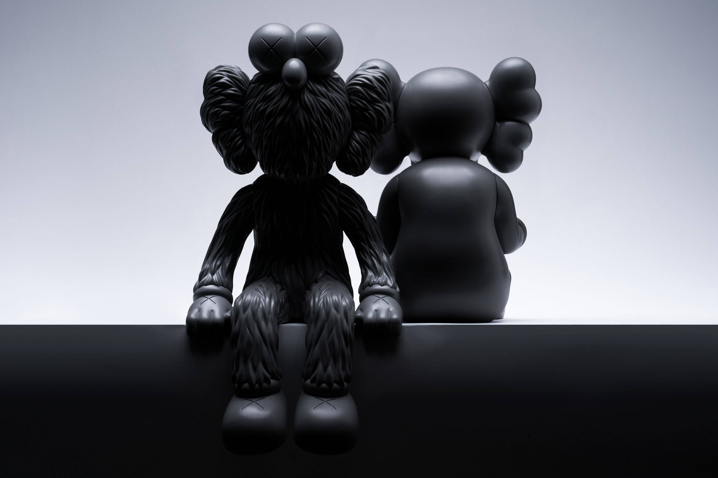 KAWS 桌布 壁紙 — 兩個啞光黑色KAWS公仔並排坐在光滑的黑色表面上，背景為漸變灰色。
