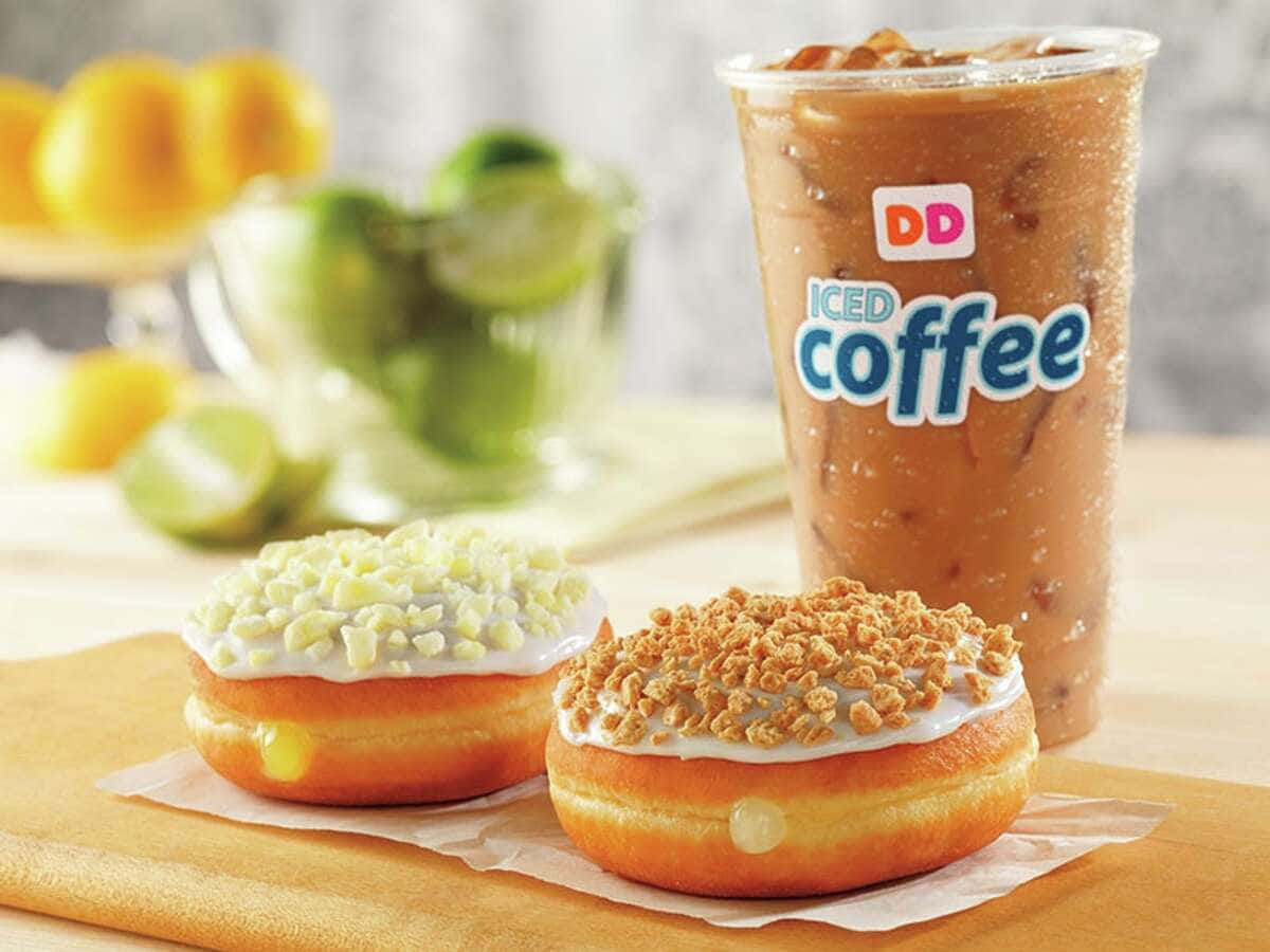 Dunkin Donuts 桌布 壁紙 — 兩個塗上白色糖霜和脆脆糖粒的釉面甜甜圈，旁邊放著一杯高身冰咖啡飲品，背景有柑橘類水果。