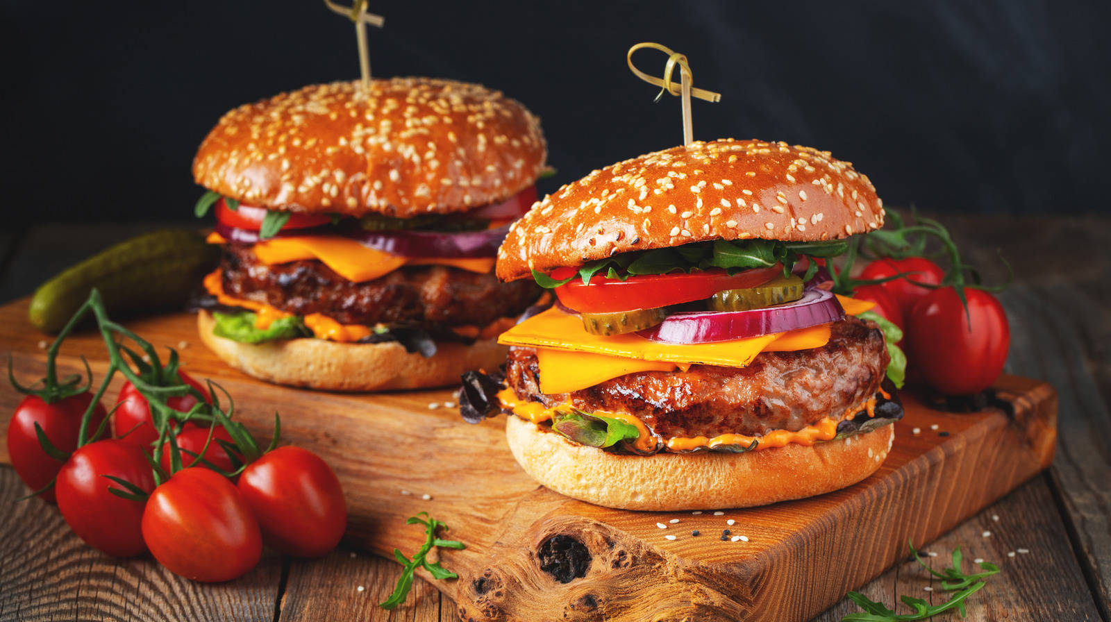 Burger King 桌布 壁紙 — 兩個多汁的芝麻籽漢堡，夾著起司、番茄和洋蔥，放在一個質樸的木板上，周圍是新鮮的櫻桃番茄和一個醃黃瓜。