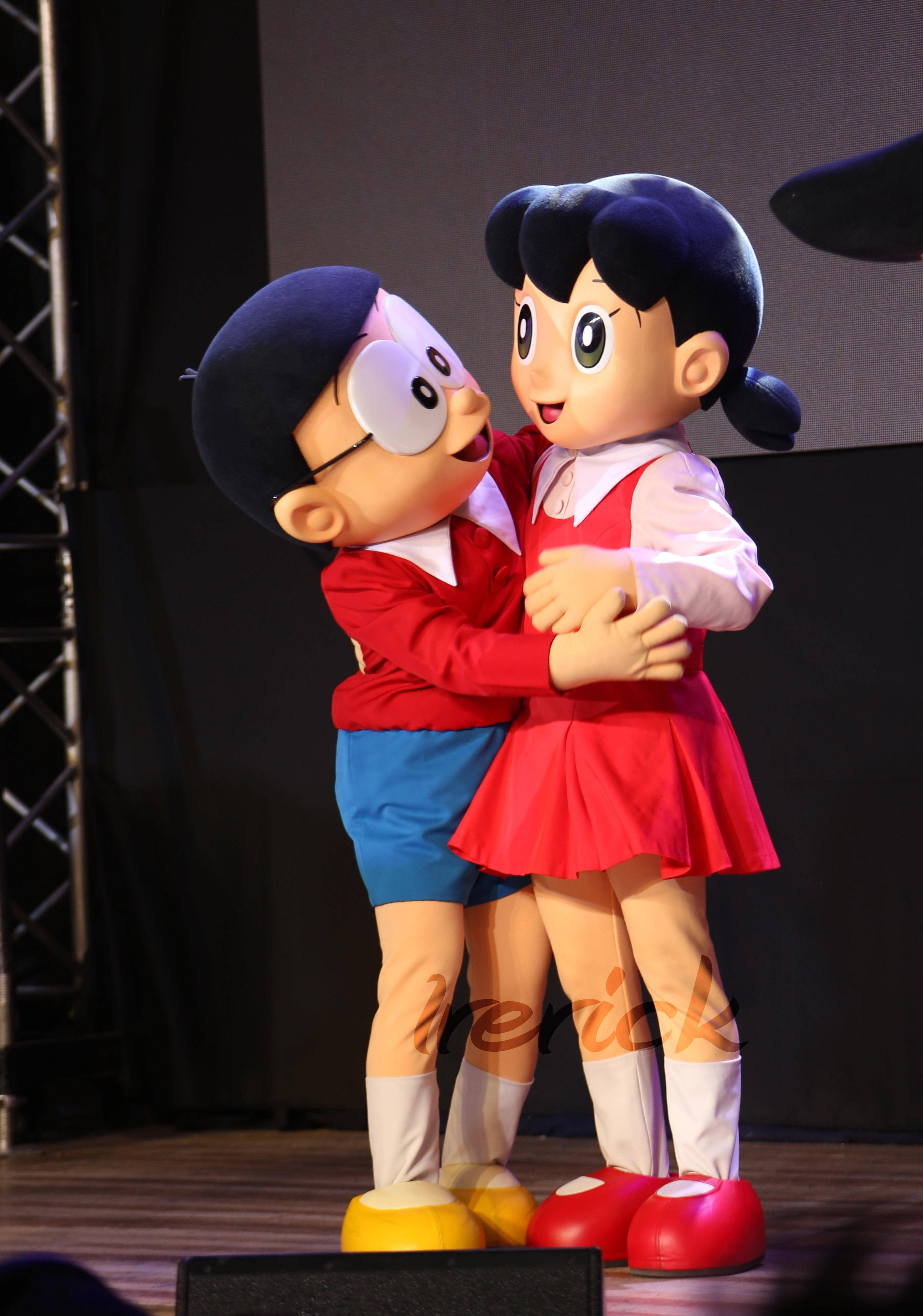 Doraemon 桌布 壁紙 — 兩個大型動漫吉祥物哆啦A夢和靜香在黑暗背景和可見燈光設備的舞台上溫暖擁抱。