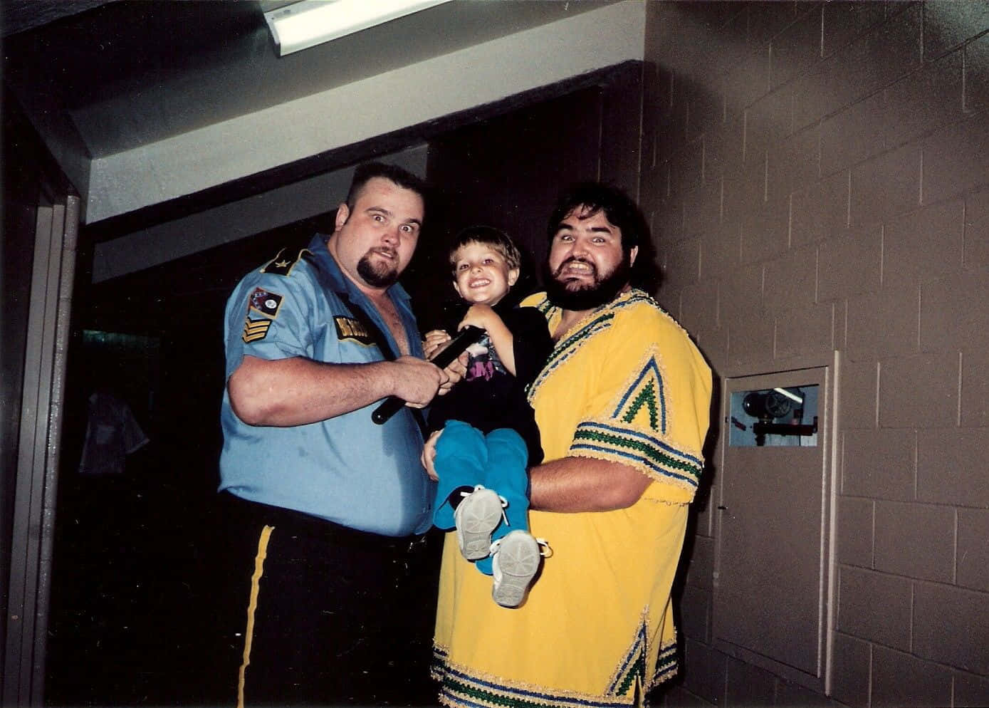 Big Boss Man 桌布 壁紙 — 兩個大男人和一個小孩在光線昏暗的走廊裡一起擺姿勢，磚牆和監視器清晰可見。