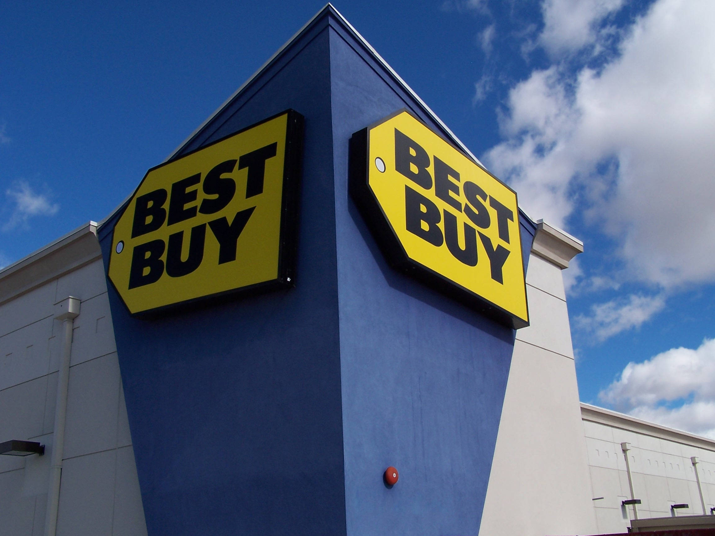buy 桌布 壁紙 — 兩個大膽的黃色Best Buy標誌安裝在藍色建築物角落，背景是明亮的藍天和蓬鬆的白雲。