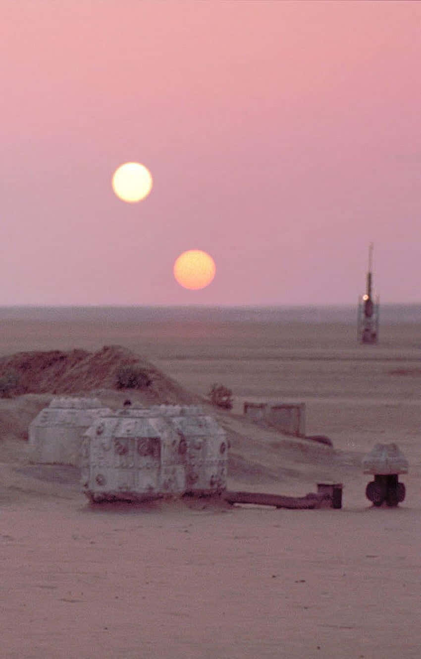 Tatooine 桌布 壁紙 — 兩個太陽在粉紅色暮色天空下的沙漠景觀中落下，遠處有未來風格的圓頂結構和一座塔。