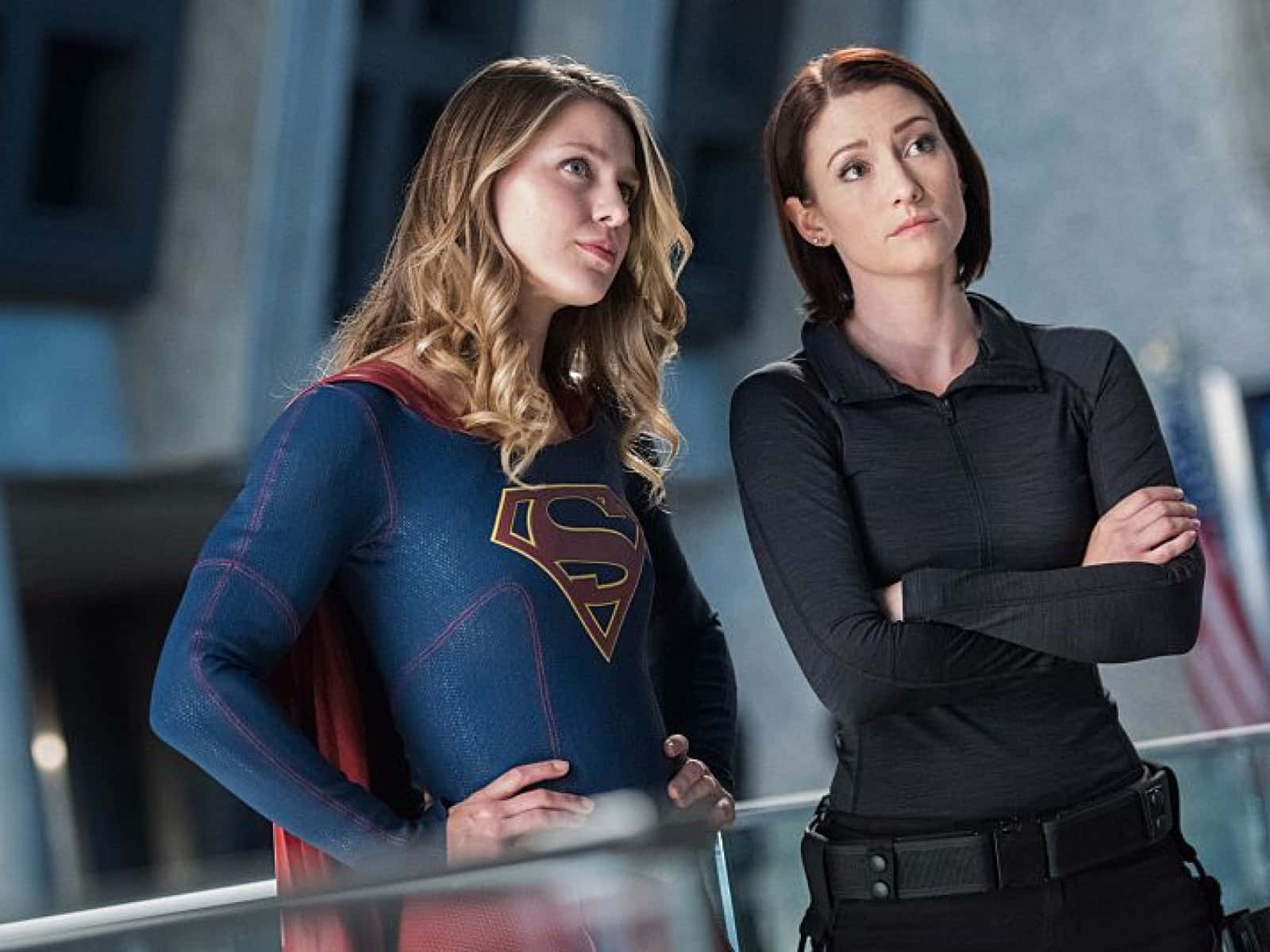Chyler Leigh 桌布 壁紙 — 兩個女人並肩而立，一個穿著帶有S標誌的藍紅色超級英雄套裝，另一個穿著黑色戰術裝備，兩人都望向遠方。
