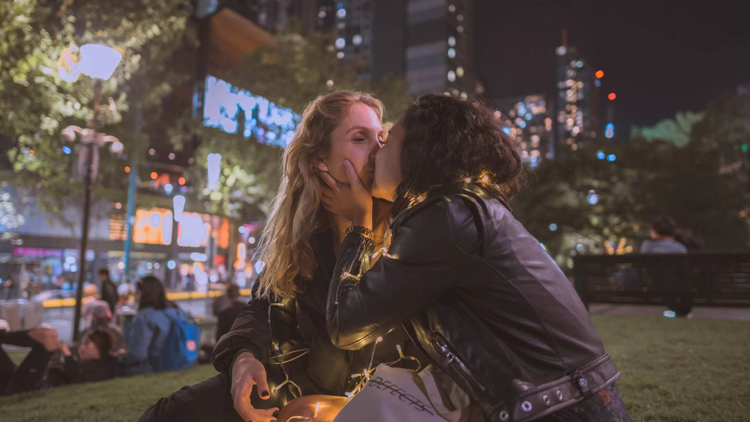 couple kiss 桌布 壁紙 — 兩個女人在夜晚的草地上城市公園中，在閃爍的城市燈光和模糊的霓虹燈牌下熱情接吻。