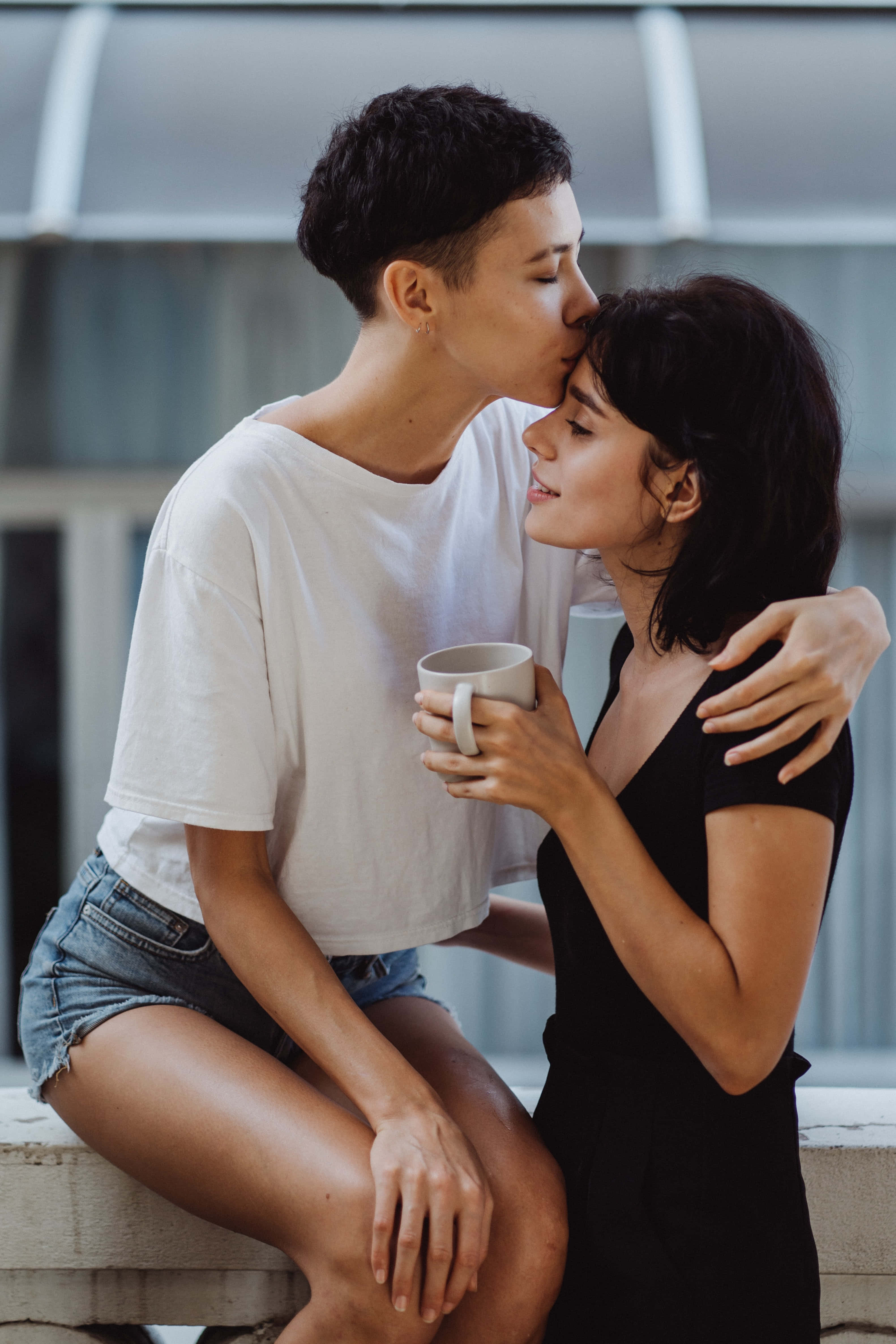 Forehead Kiss 桌布 壁紙 — 兩個女人在屋頂上分享親密時刻，一個親吻另一個的臉頰，同時拿著一個杯子。