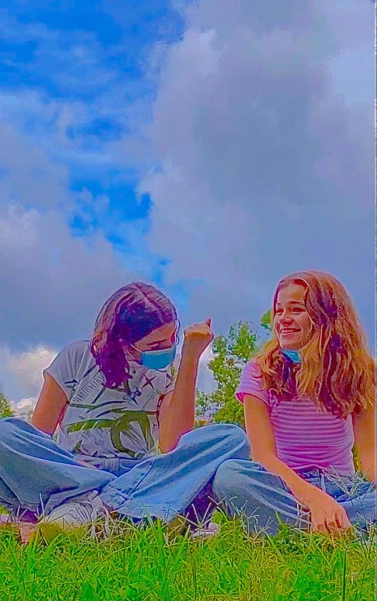 獨立美學動圖 桌布 壁紙 — 兩個女孩坐在鮮綠色的草地上，在明亮的藍天和蓬鬆的白雲下戴著口罩微笑。