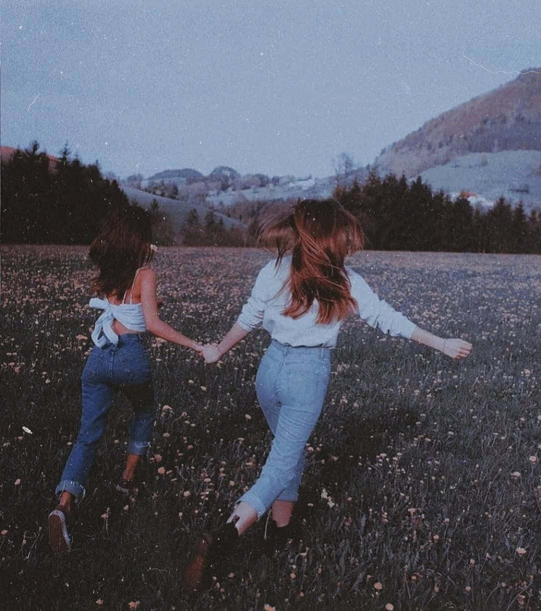 Best Friend Aesthetic 桌布 壁紙 — 兩個女孩手牽手跑過一片野花田，暮色天空下遠山依稀可見。