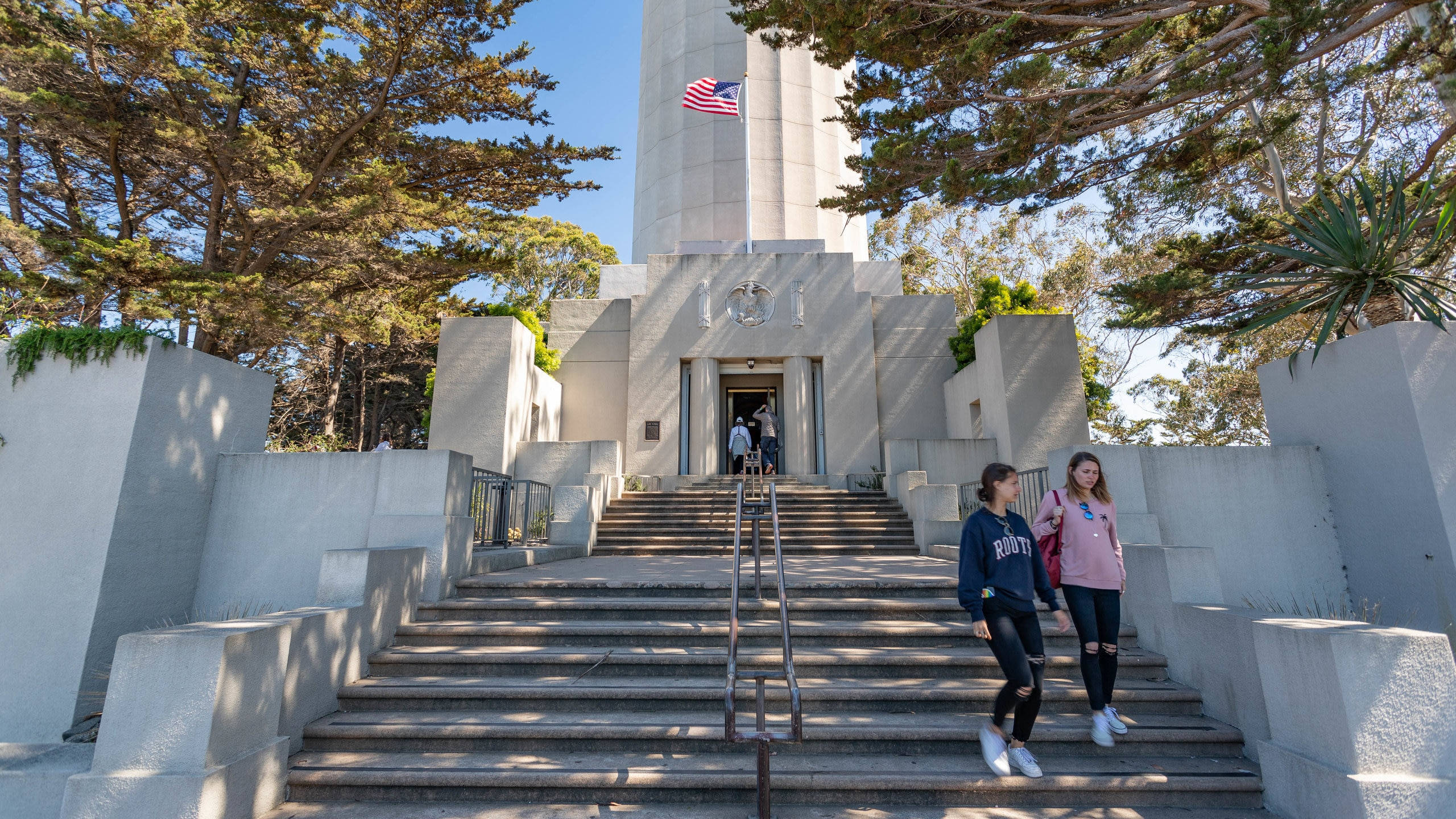 San Francisco 桌布 壁紙 — 兩個女孩沿著石階走向一座高大的白色紀念碑，頂端有美國國旗，周圍是高聳的樹木，陽光明媚。