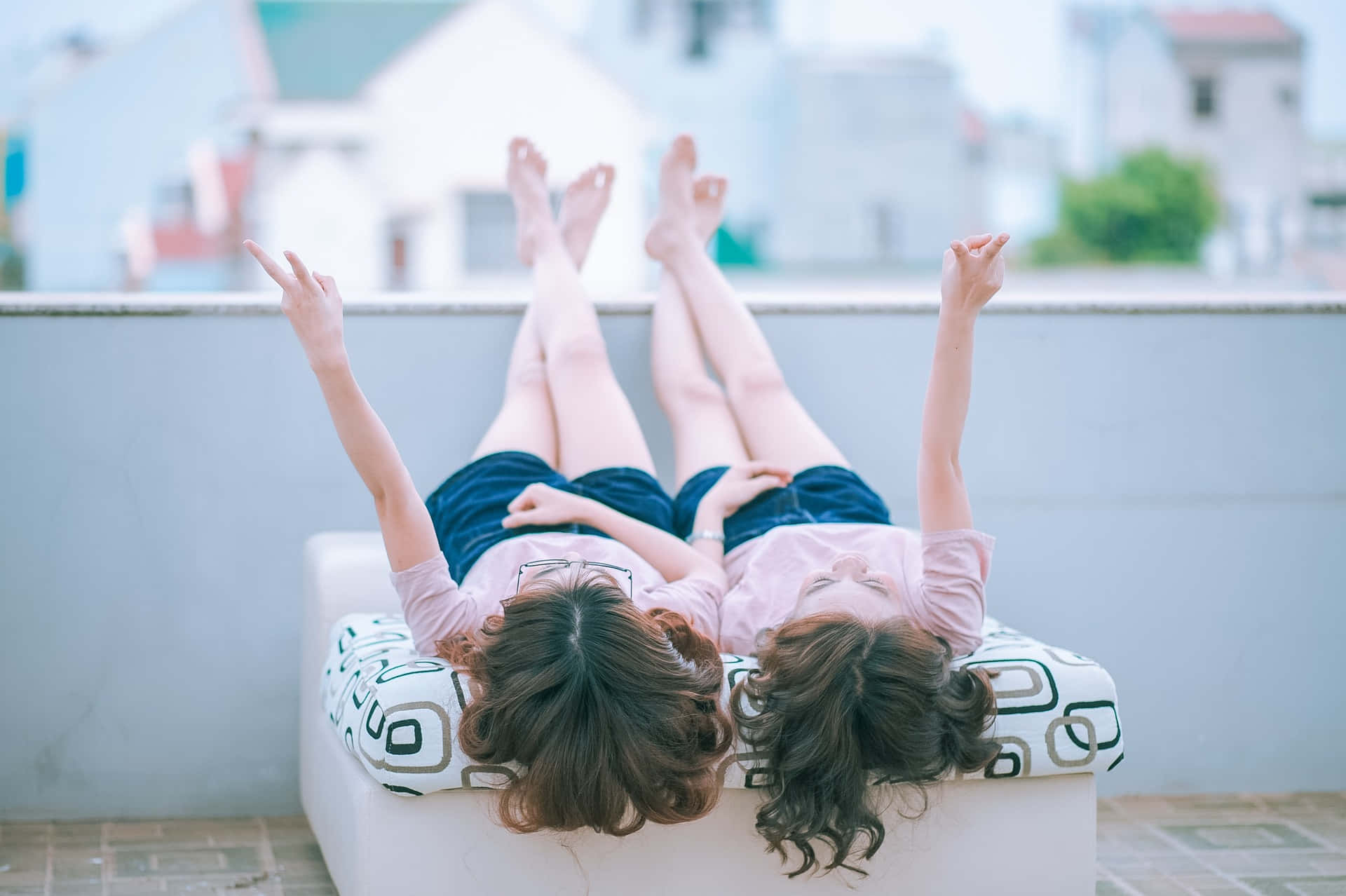 女同志 桌布 壁紙 — 兩個女孩躺在天台沙發上，雙腿抬起，雙臂高舉，在柔和模糊的城市背景下盡情歡樂。