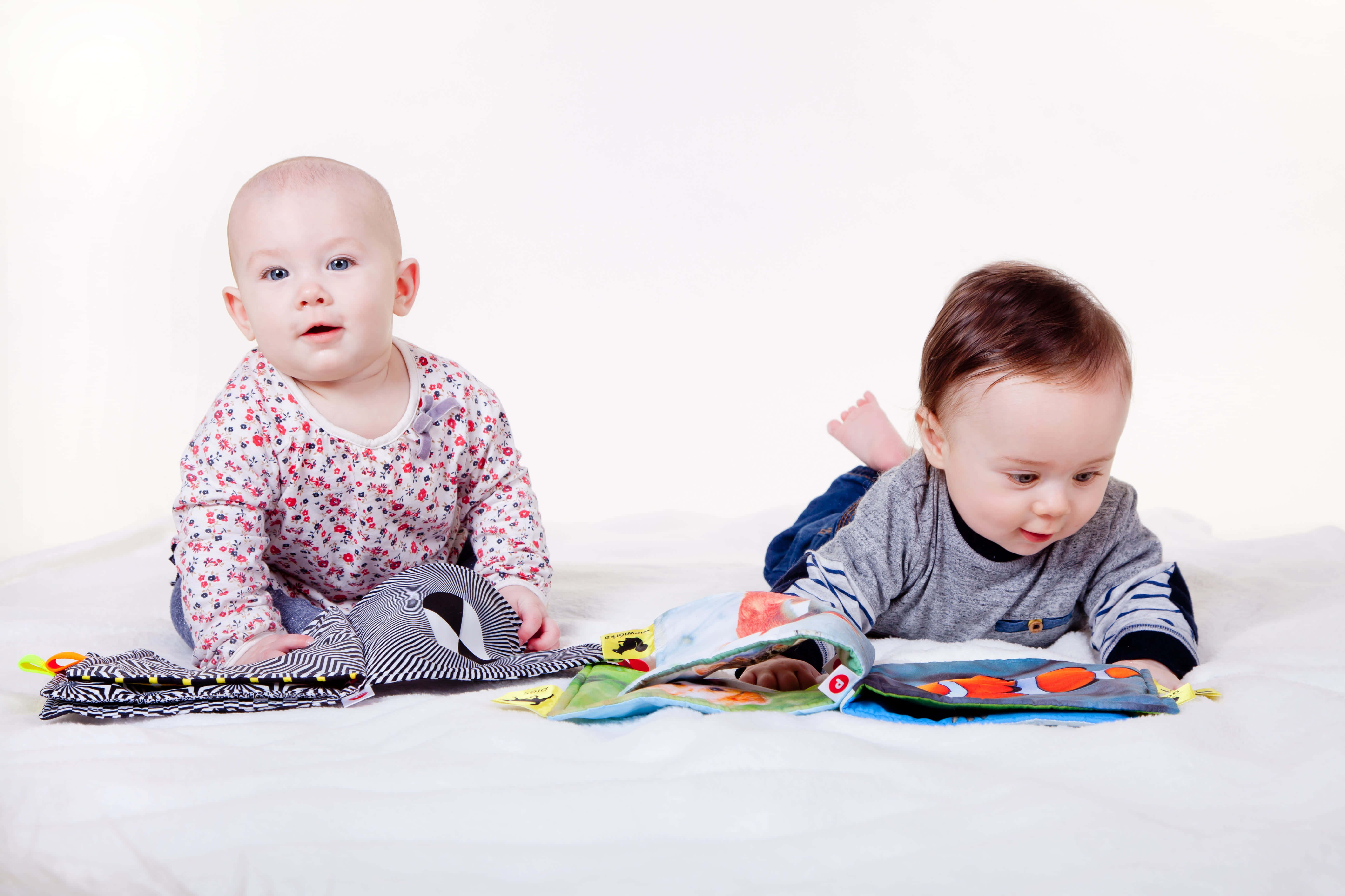 Cute Babies 桌布 壁紙 — 兩個好奇的嬰兒躺在白色床上，探索色彩繽紛、圖案明亮的紋理書。