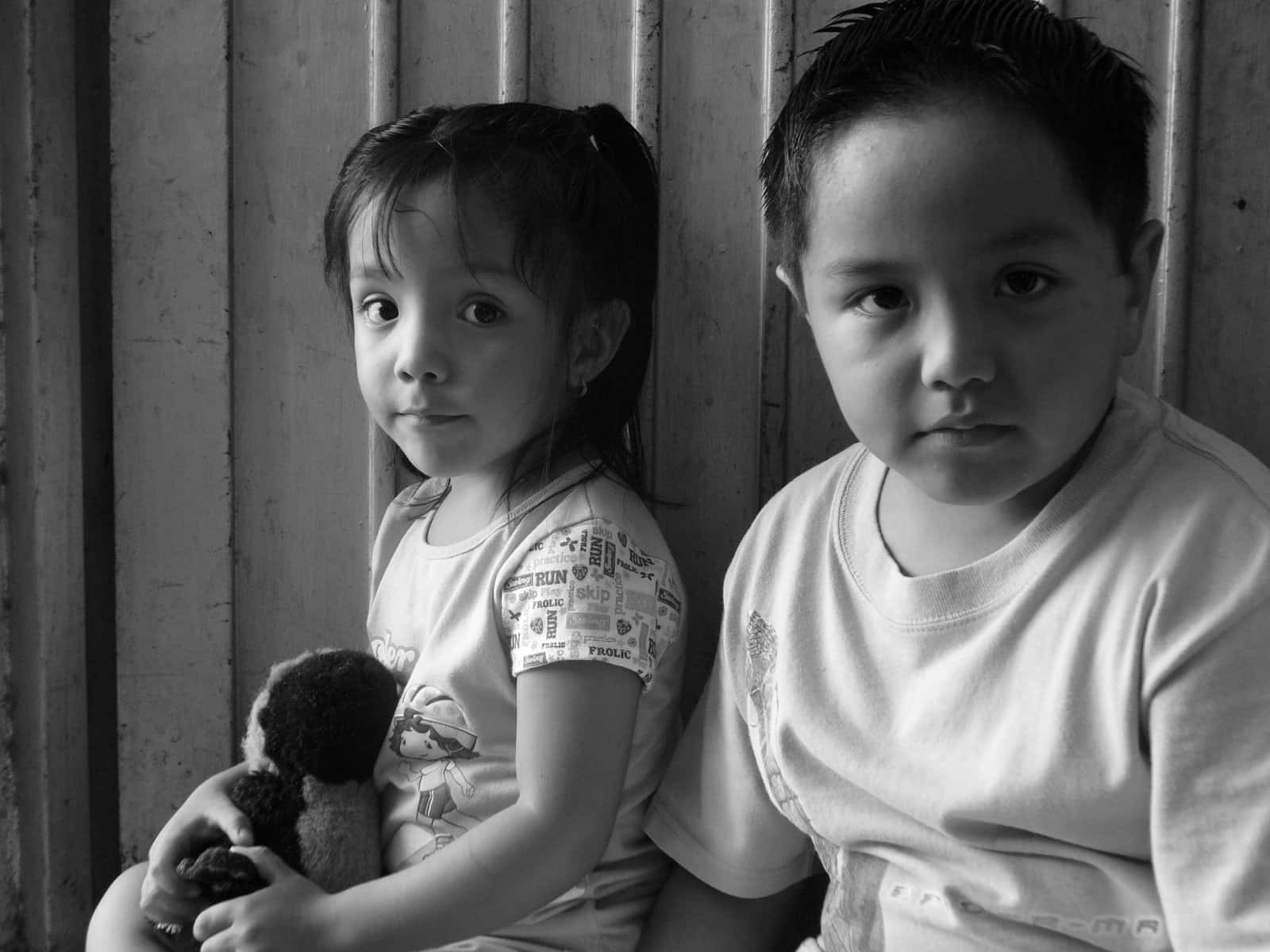 Brother And Sister 桌布 壁紙 — 兩個孩子並排坐在木牆旁，女孩抱著一個毛絨玩具，兩人都直視鏡頭，形成一幅黑白肖像。