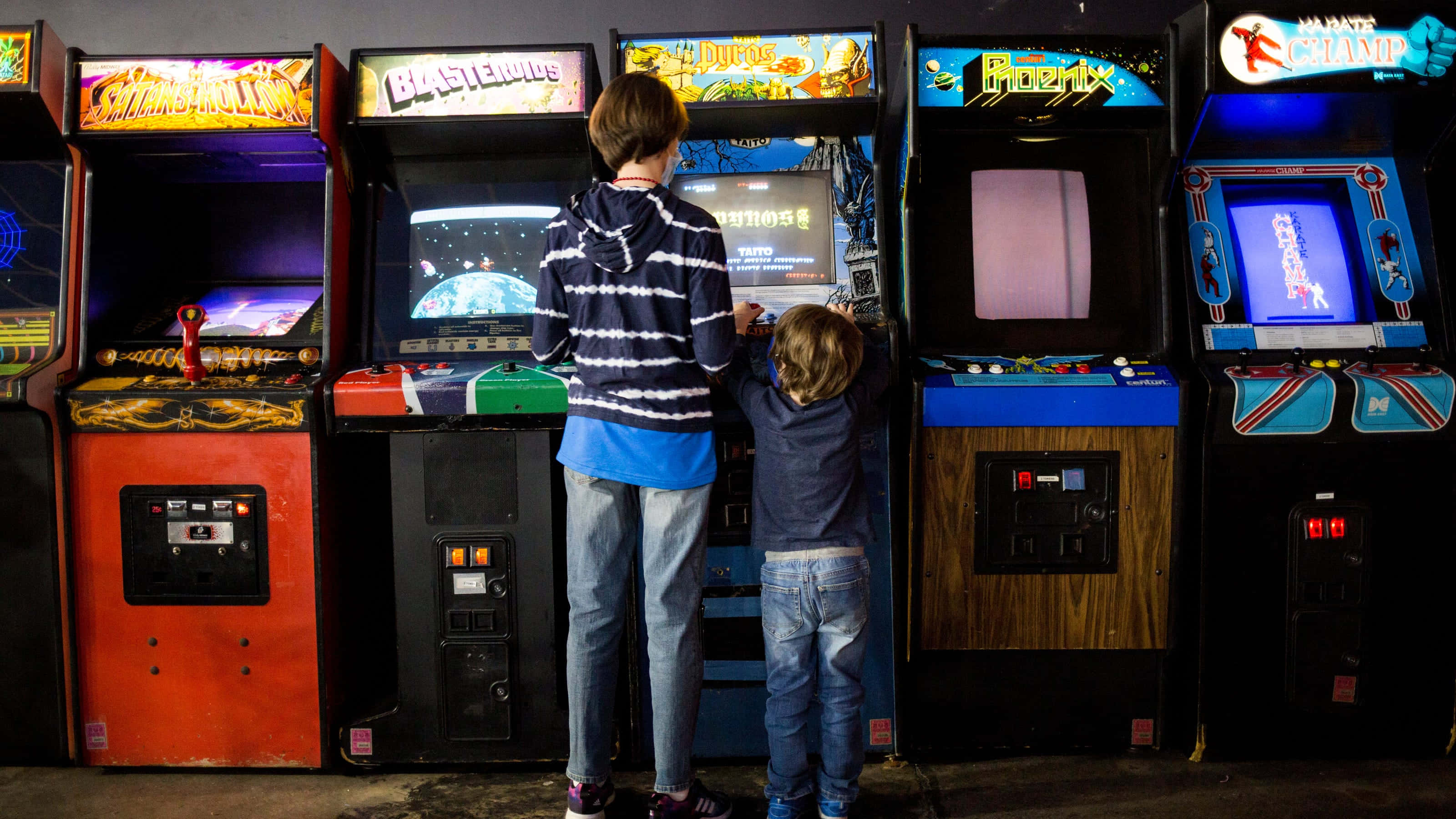 Arcade Games 桌布 壁紙 — 兩個孩子並肩站在一條燈光昏暗的復古遊戲巷裡，全神貫注於閃爍著彩色燈光的懷舊街機遊戲。
