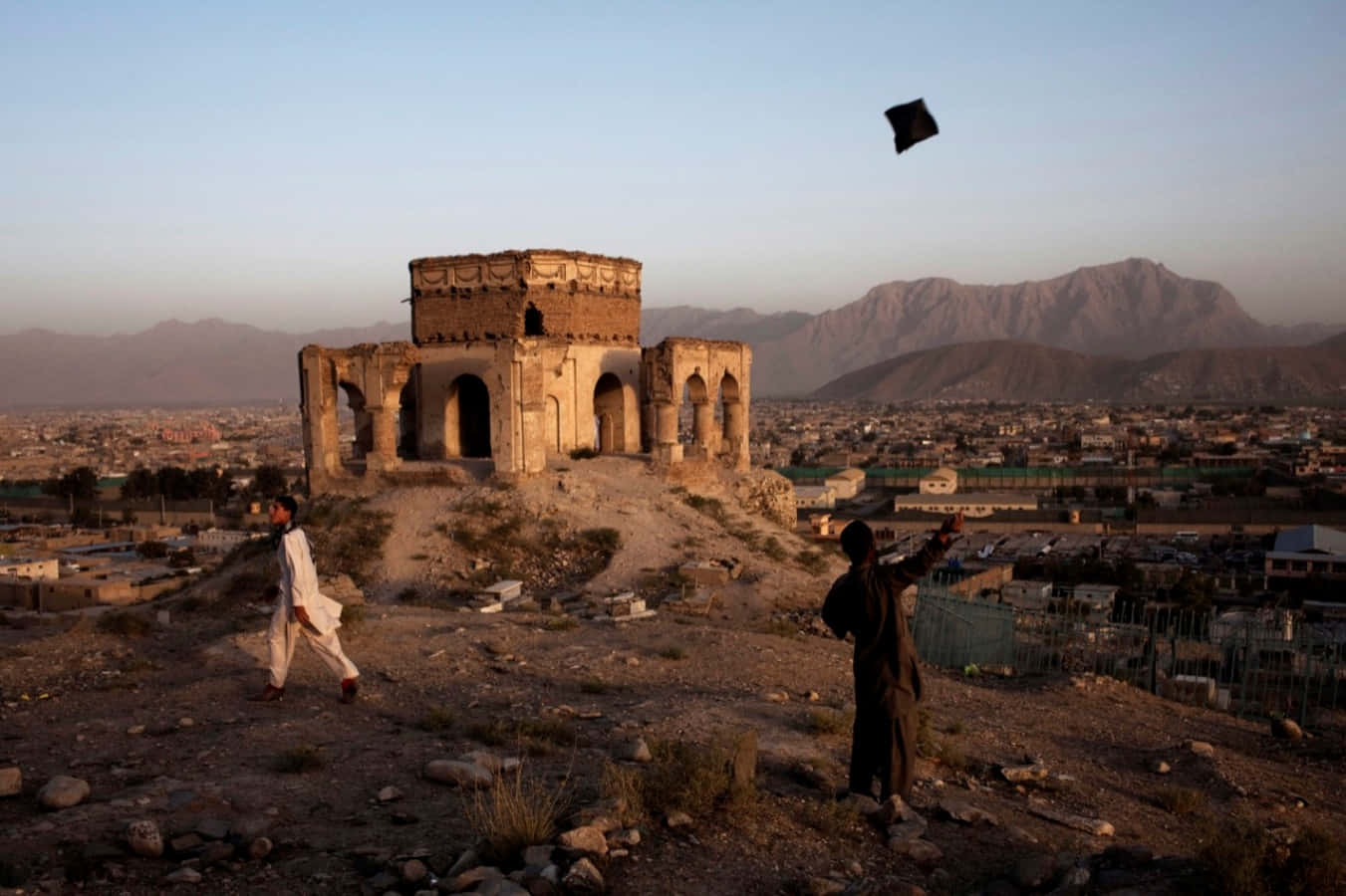 Afghanistan 桌布 壁紙 — 兩個孩子在一個風化的古老廢墟附近放風箏，俯瞰著黃金時段中一座廣闊的城市。