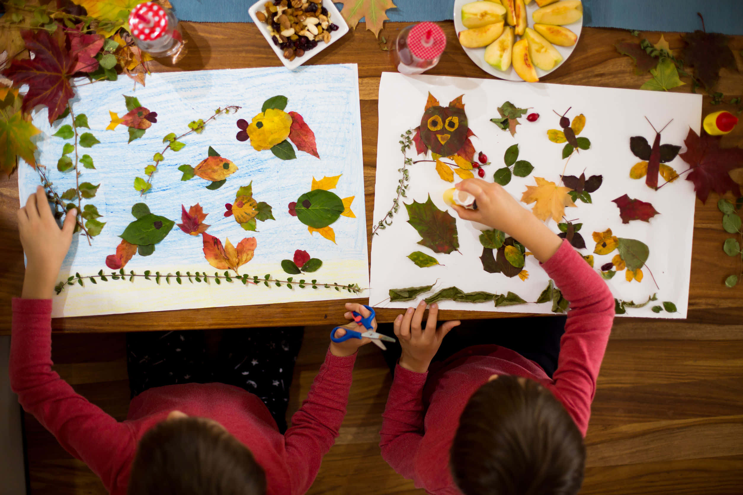 Fall Crafts 桌布 壁紙 — 兩個孩子在木桌上用紙創作秋季樹葉藝術，周圍環繞著季節性樹葉和小吃。