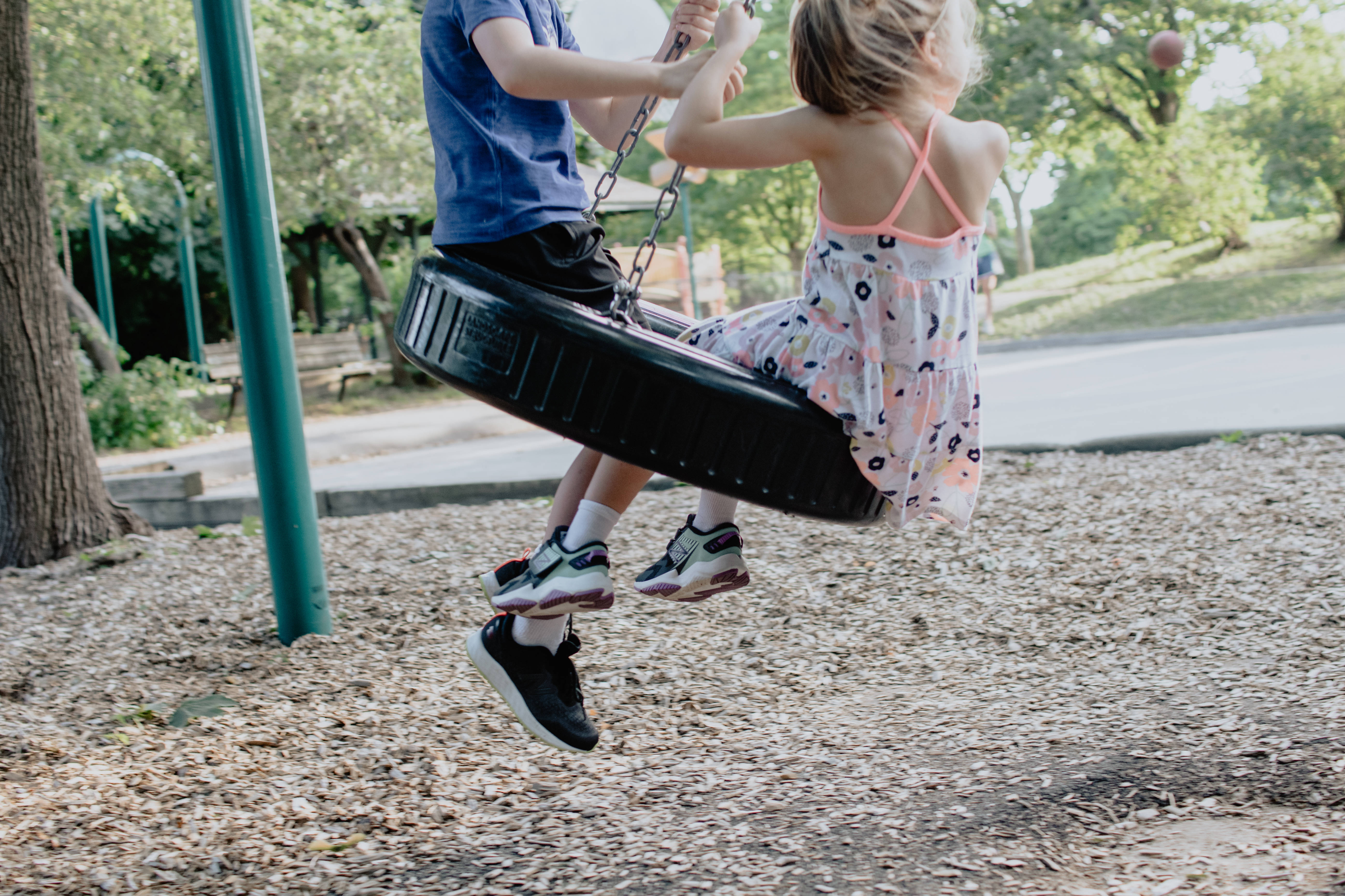 Playground 桌布 壁紙 — 兩個孩子在陽光明媚的公園裡一起盪著輪胎鞦韆，笑著，下面有樹木和覆蓋物。