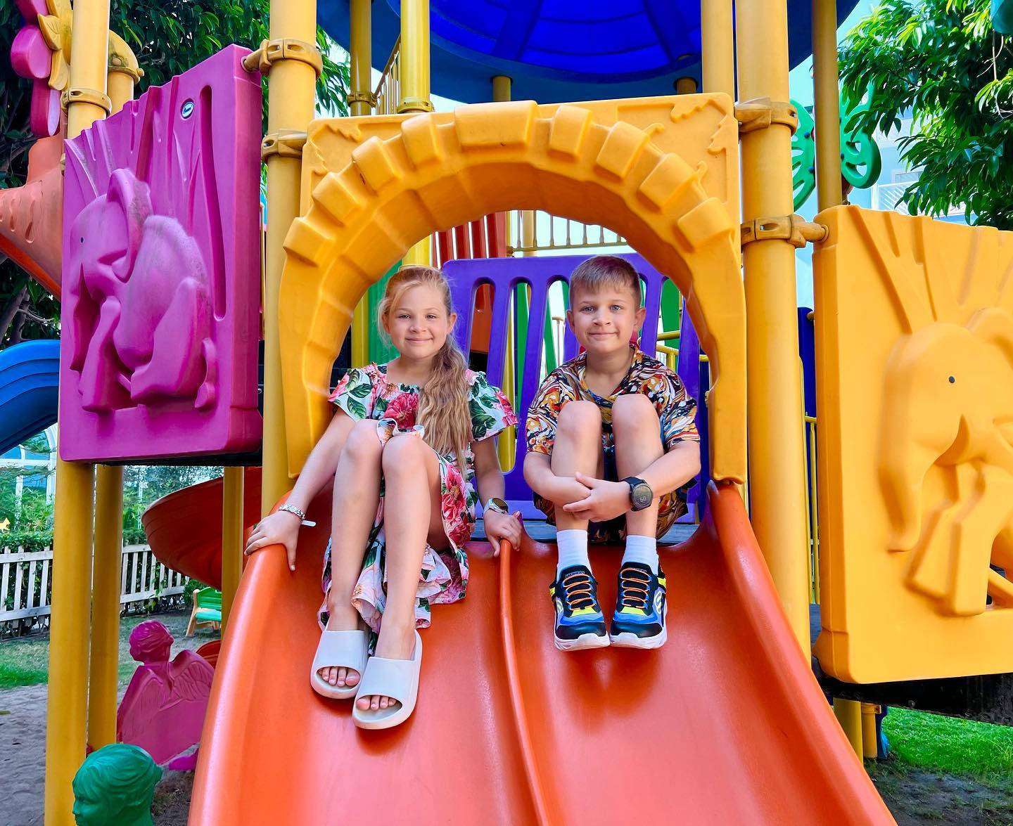 Playground 桌布 壁紙 — 兩個孩子坐在一個色彩繽紛的遊樂場結構上，一個亮橙色的溜滑梯頂端，在晴朗的日光下。
