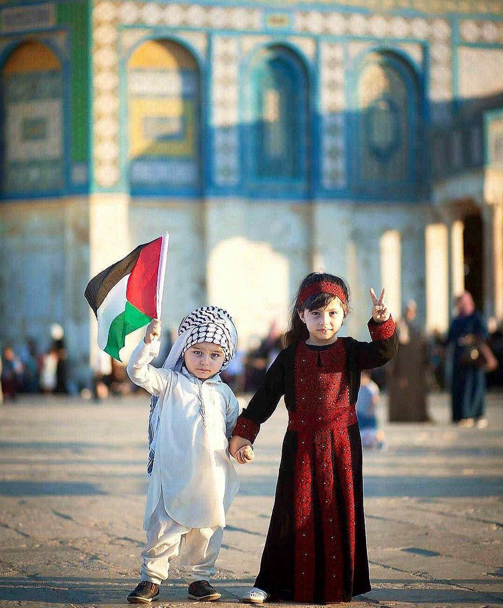 Palestine 桌布 壁紙 — 兩個孩子手牽手，一個揮舞巴勒斯坦旗幟，另一個做出和平手勢，背景是一座色彩繽紛、裝飾華麗的建築物。